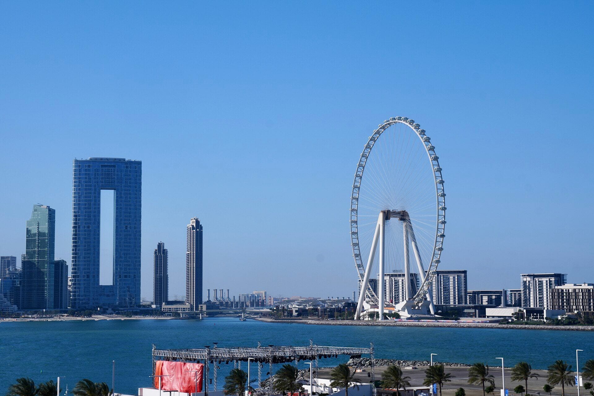 The white Ain Dubai observation wheel on Bluewaters Island beside the twin towers of Address Beach Resort under a clear blue sky in Dubai