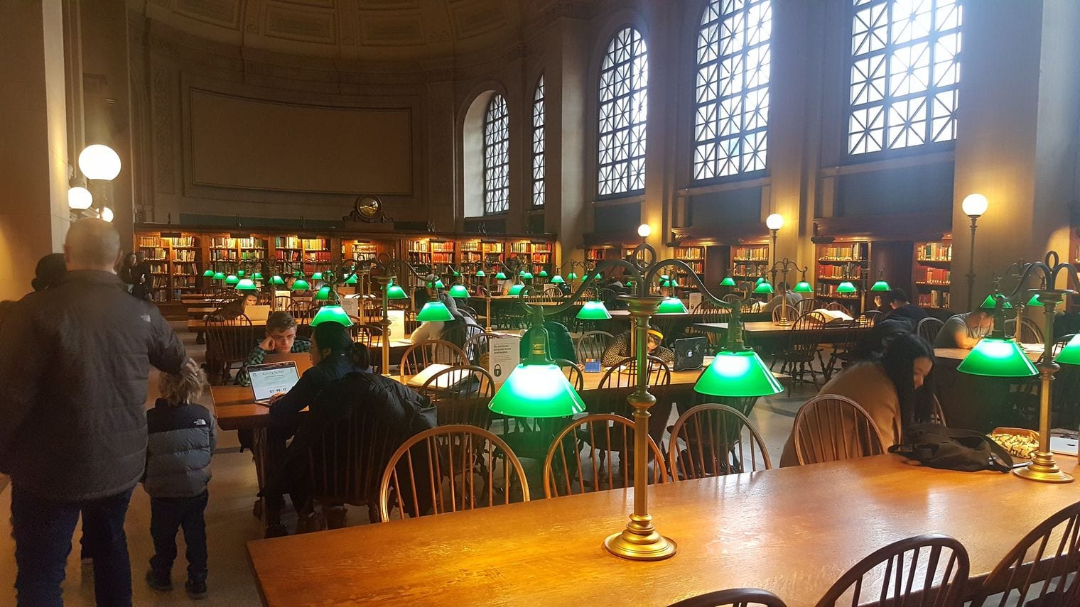 Readers at long wooden tables under rows of green banker's lamps in the barrel-vaulted Bates Hall reading room at Boston Public Library
