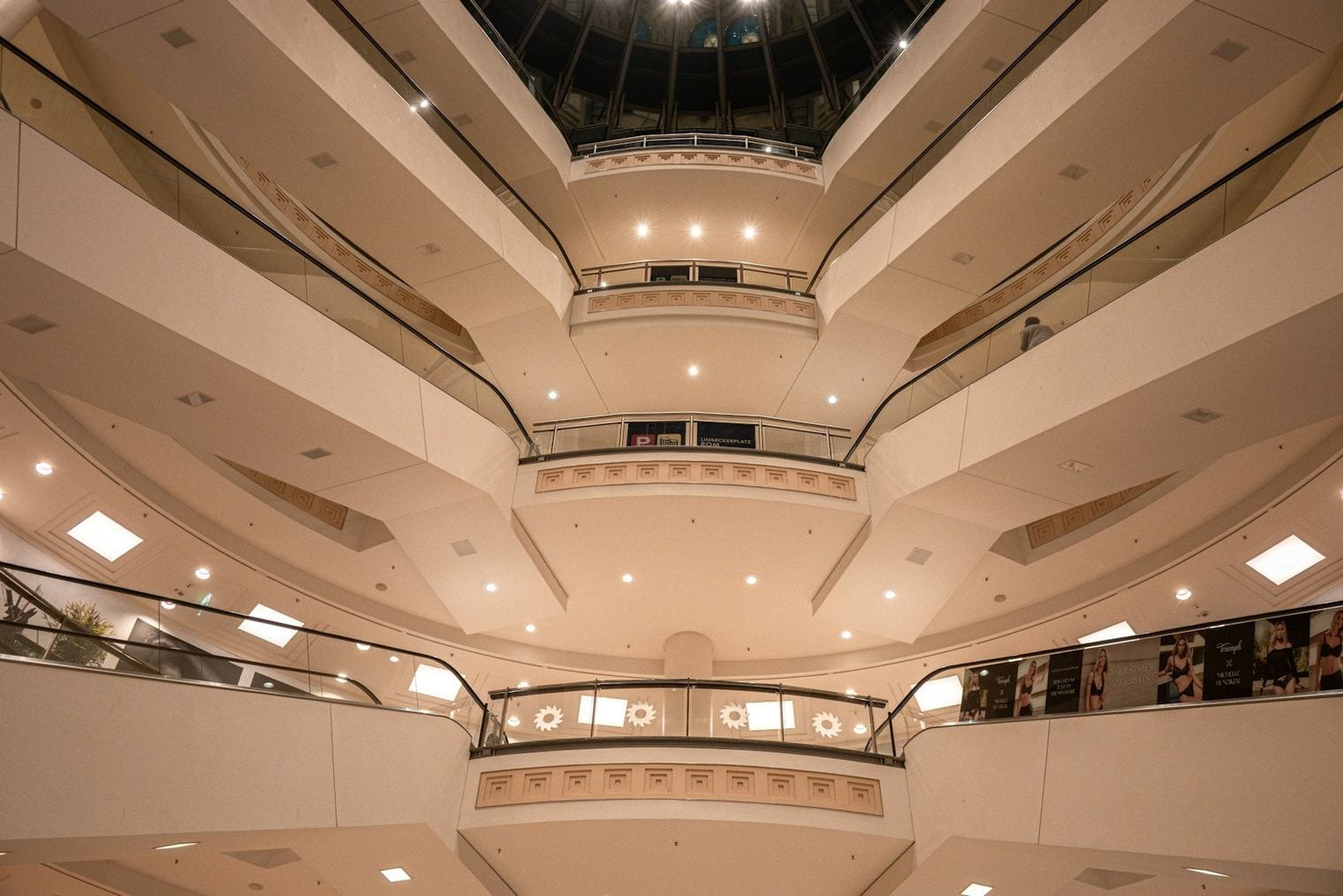 A curved multi-level shopping mall atrium, escalators rising between tiers of balconies beneath a domed skylight