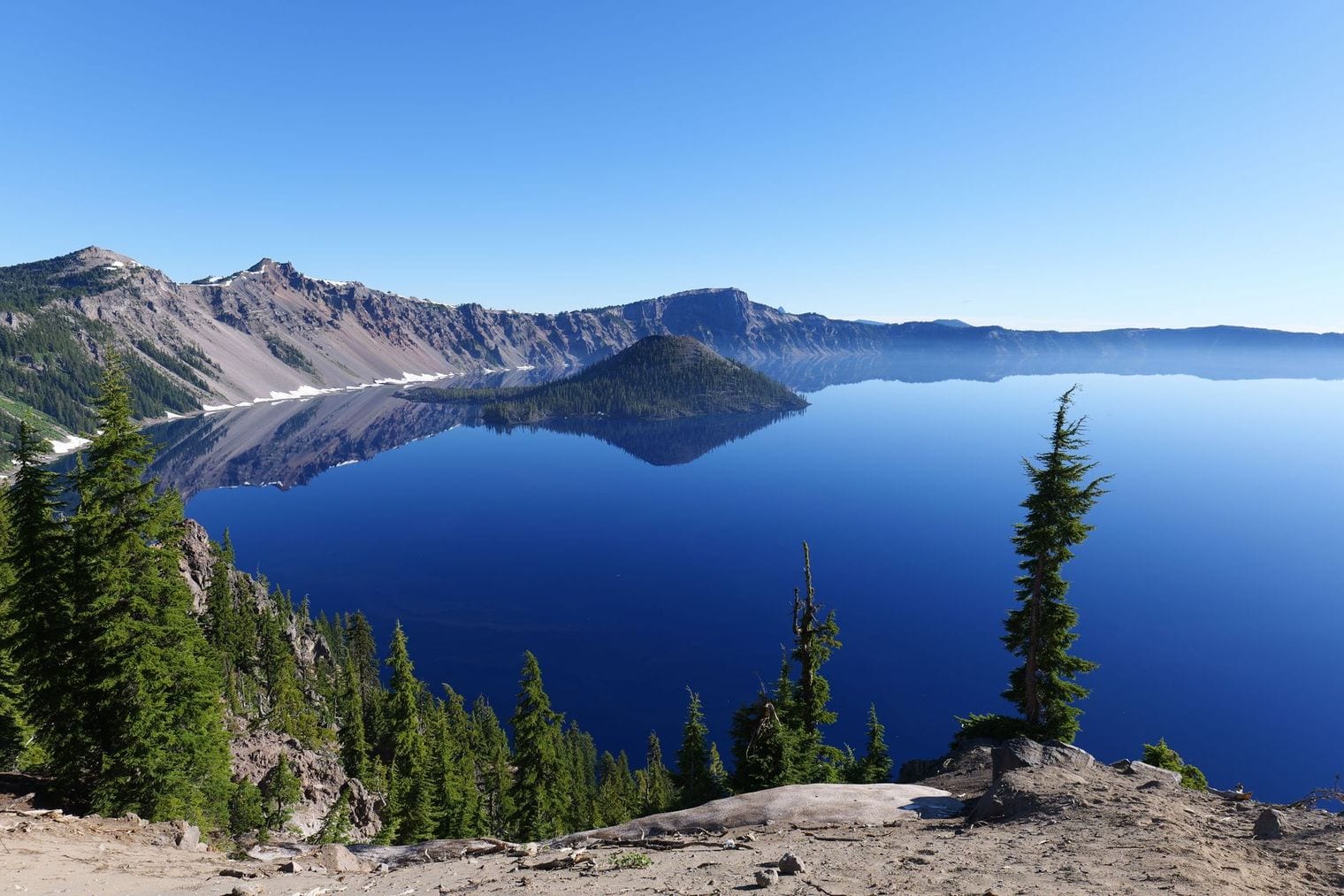 Deep blue water fills a volcanic caldera, with the forested cone of Wizard Island rising near the far shore and steep grey crater walls reflected in the still surface.