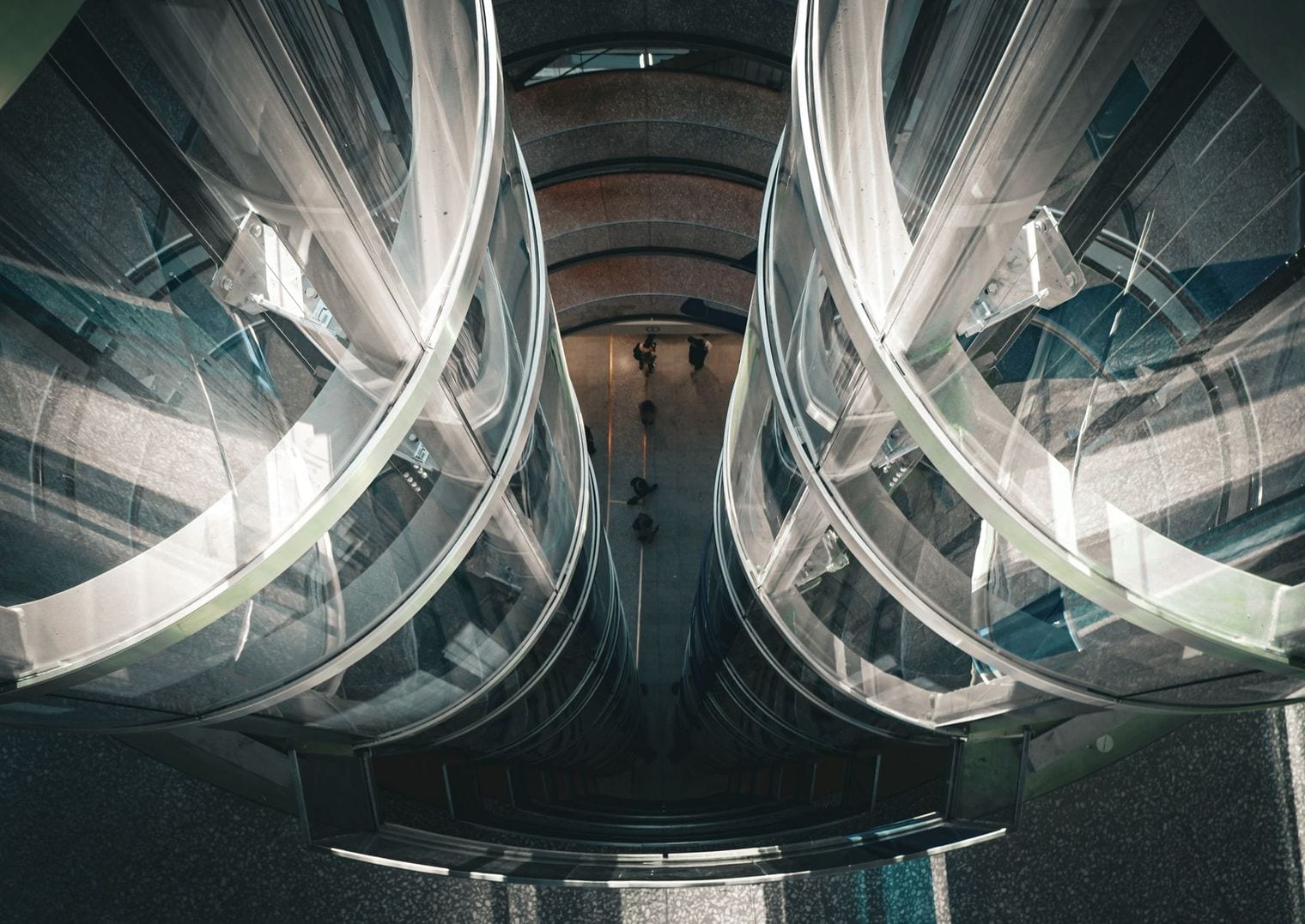 Looking down between twin glass elevator shafts in a high-rise atrium, with people small on the floor far below