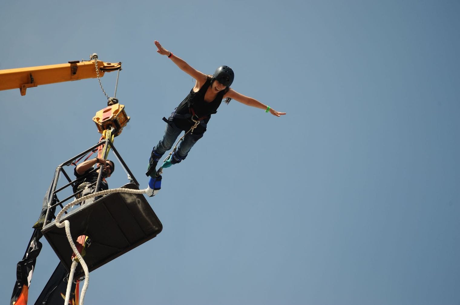 A bungee jumper leaps arms-outstretched from a crane-suspended cage against a clear blue sky, the cord trailing from her ankles as an operator watches from the platform.