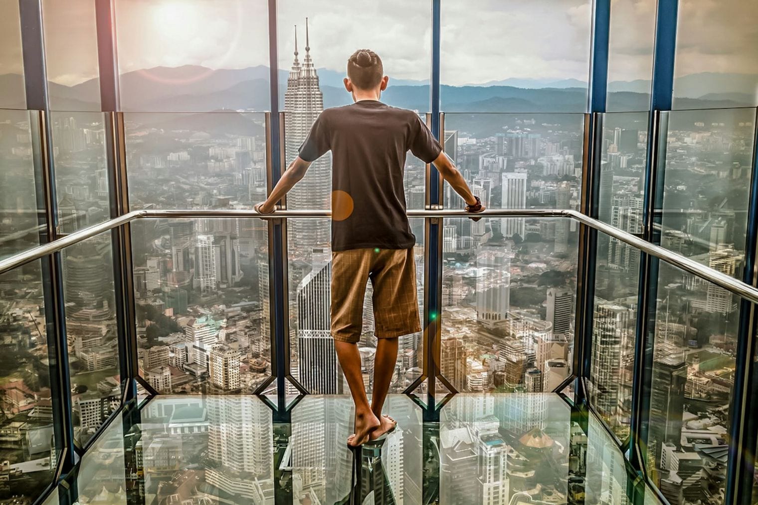 A visitor standing barefoot on a glass observation floor high above a city skyline, twin towers rising through the haze beyond