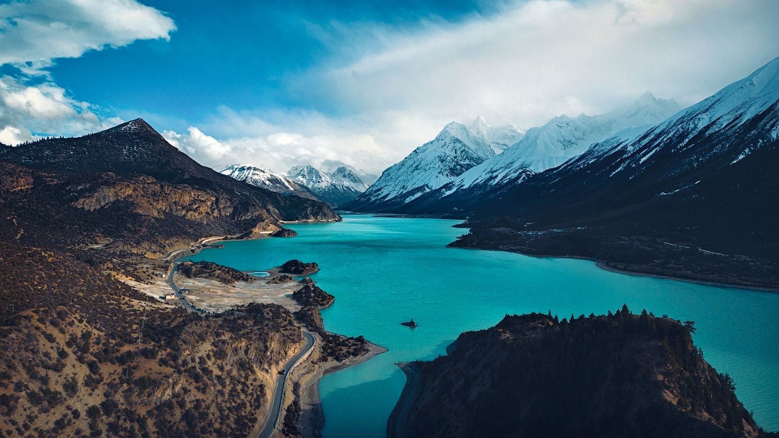 Aerial view of a turquoise high-altitude lake winding between barren brown ridges and snow-capped peaks under a clouded sky