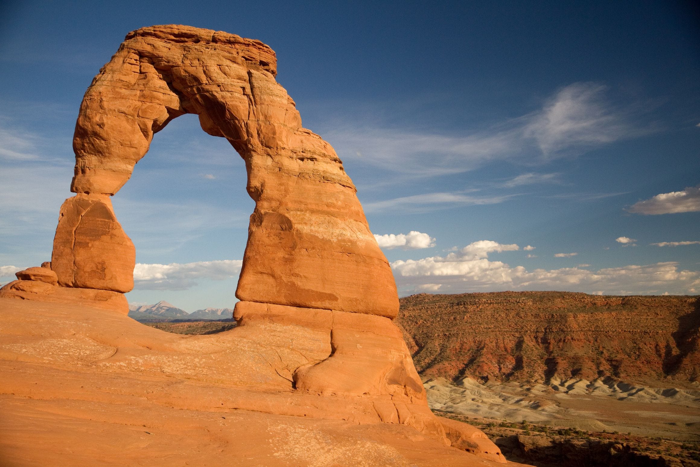 Delicate Arch standing on its slickrock bowl at golden hour, with the snow-streaked La Sal Mountains on the horizon behind it
