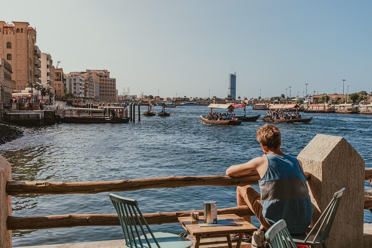 Wooden abra water taxis crossing Dubai Creek on a sunny day, seen past a man sitting at a waterfront café table in the old city.