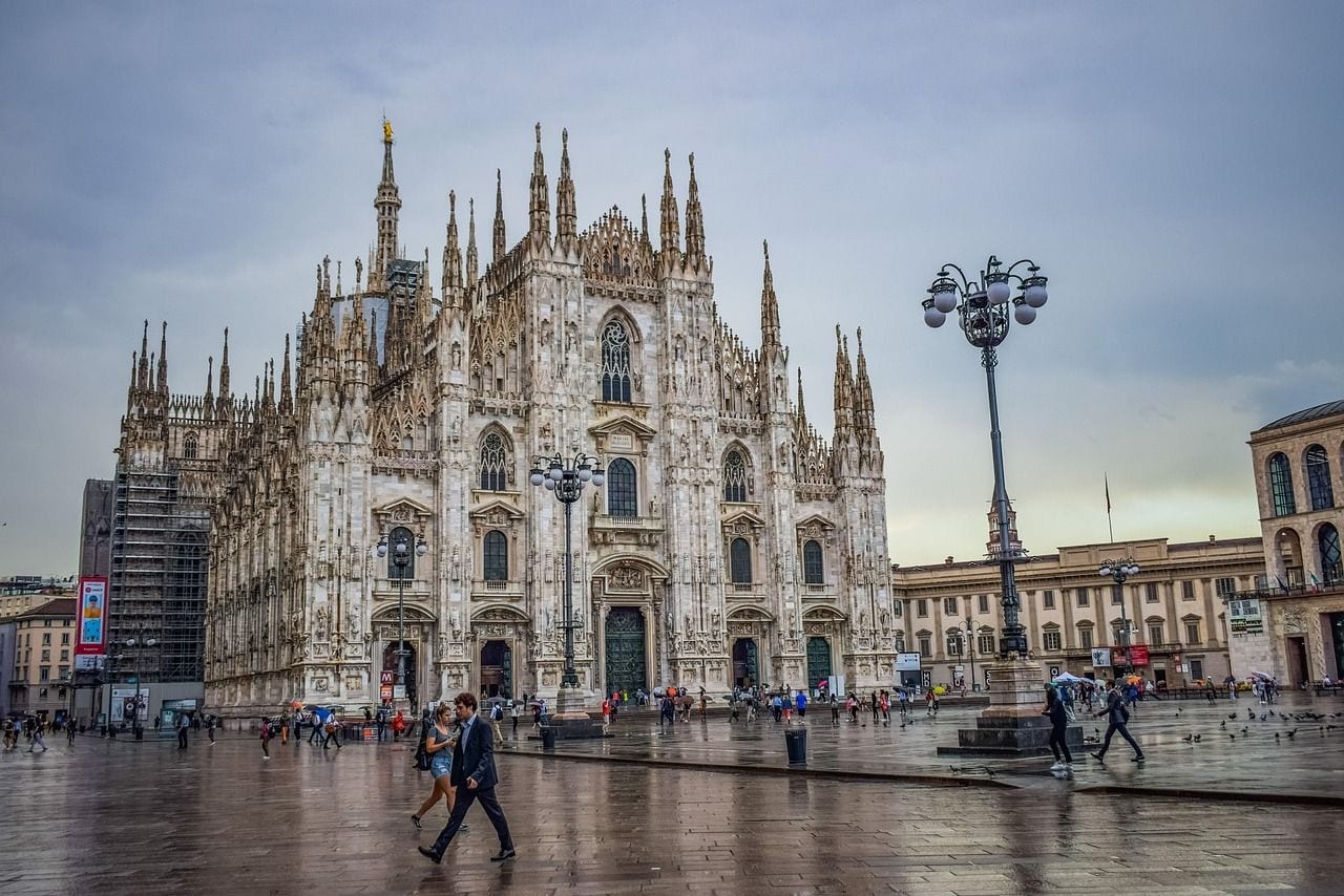 The white marble facade and spires of Milan Cathedral rising above a rain-wet Piazza del Duomo under a dramatic sky