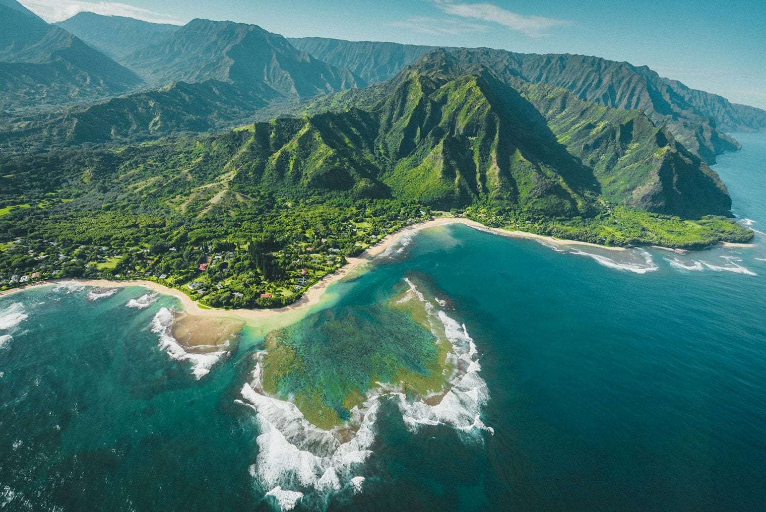 Aerial view of a turquoise bay opening to the sea between two low, densely forested headlands under a bright blue sky.