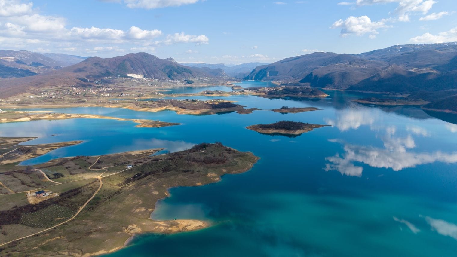 Aerial view of a turquoise lake winding between brown mountain ridges, with small wooded islands and clouds mirrored on the still surface.