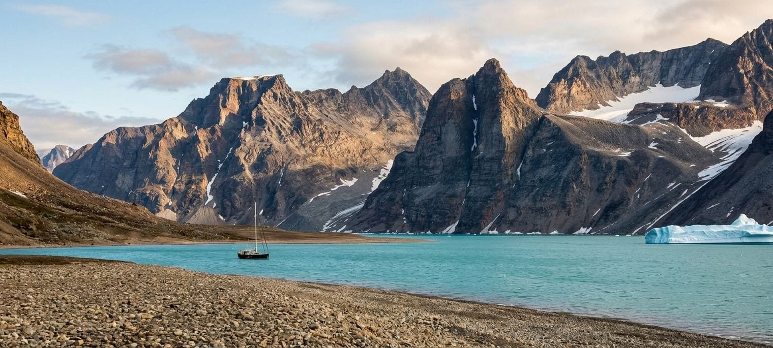 A sailing boat anchored in a calm turquoise Arctic fjord below steep snow-streaked peaks, with an iceberg drifting near the far shore