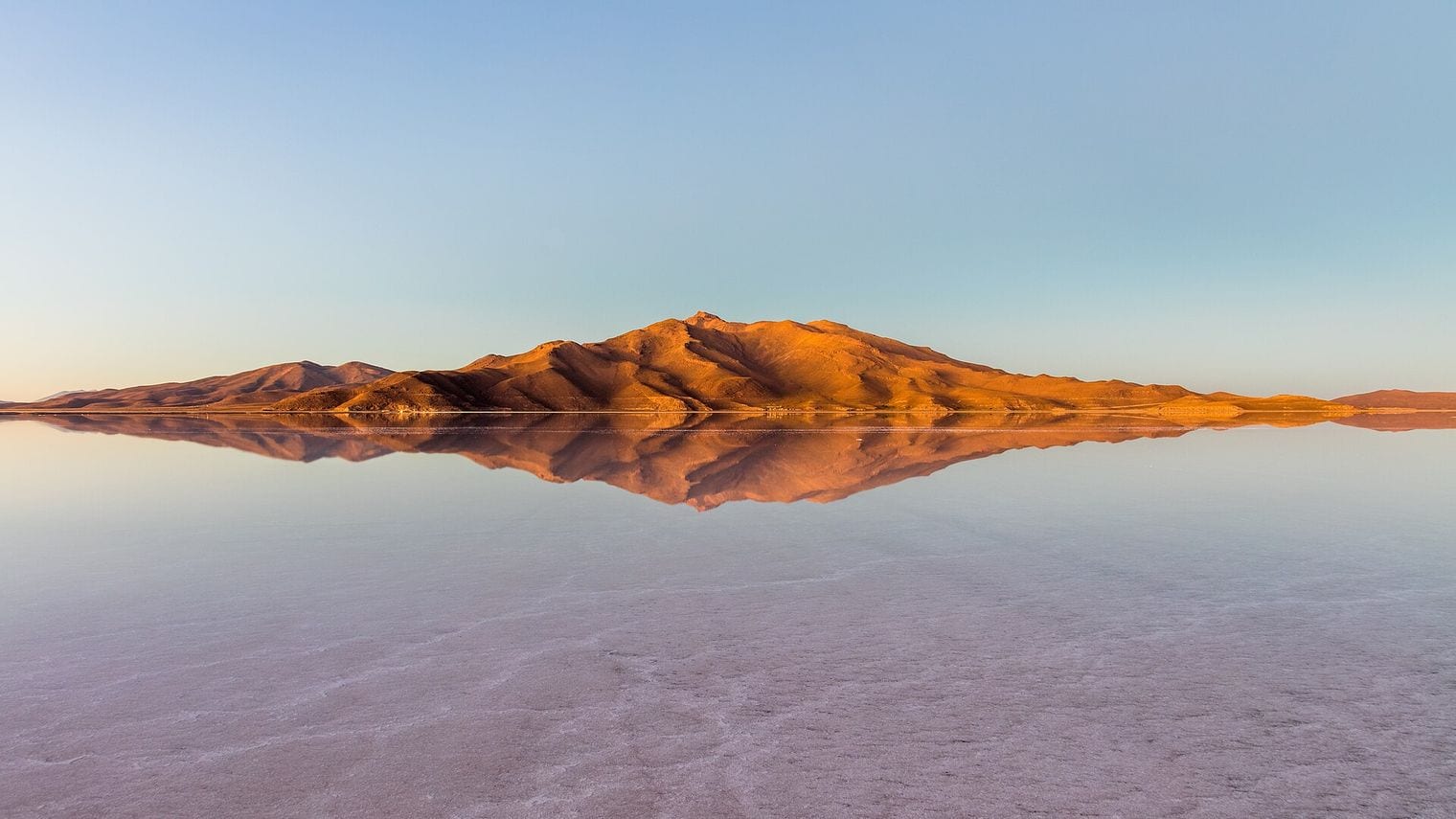A lone ridge on the Salar de Uyuni glowing orange at sunrise, mirrored in the thin water covering the salt flat