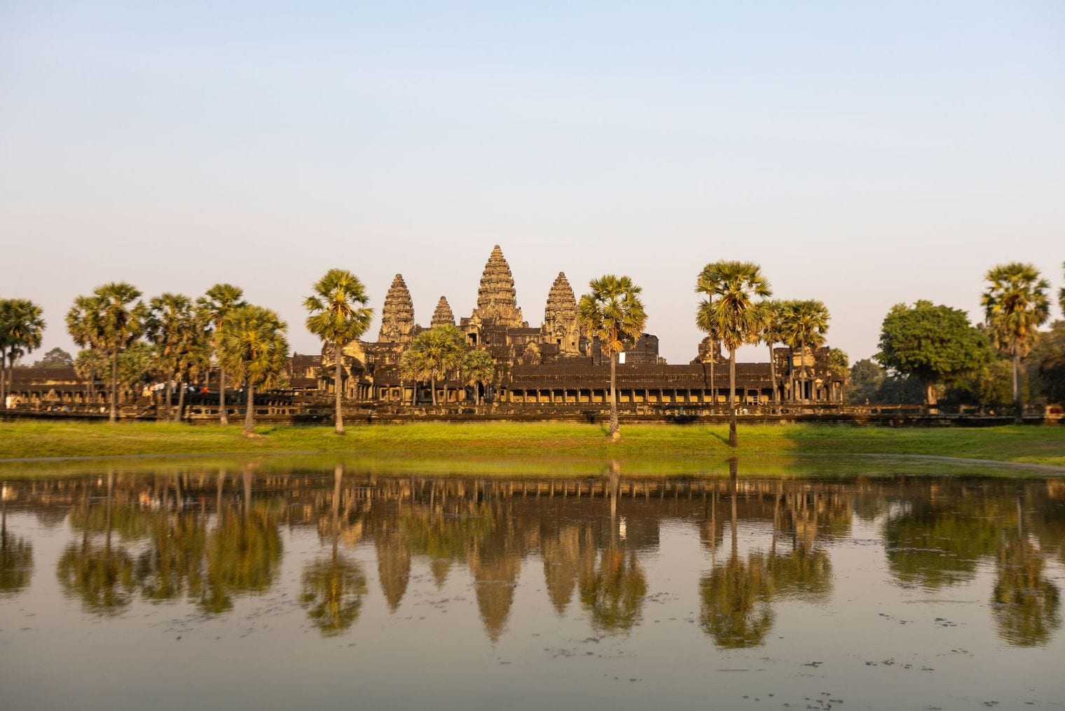 The long sandstone outer gallery of Angkor Wat mirrored in the still water of its moat at first light, with the five central towers rising behind the treeline
