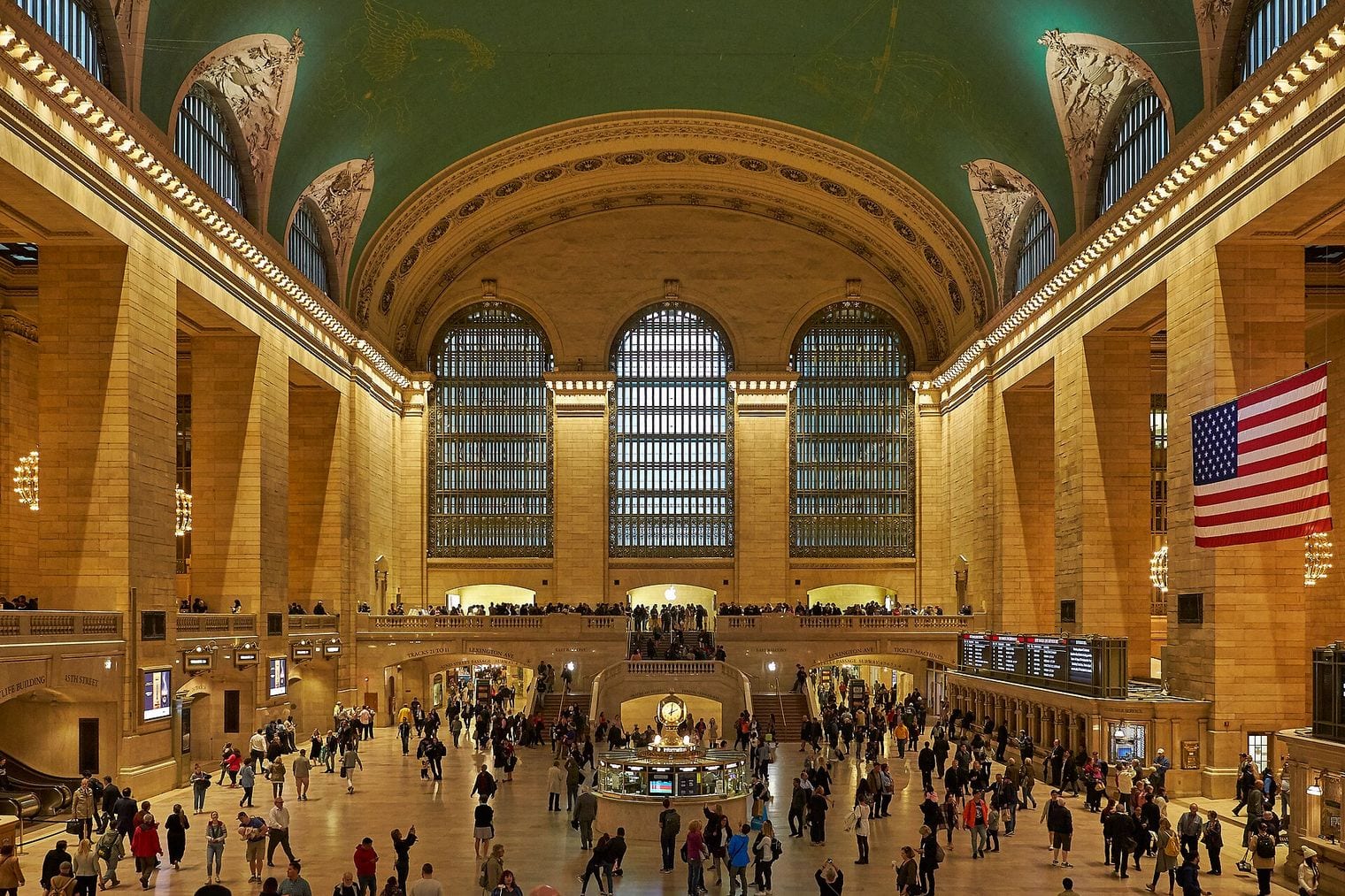 Crowds crossing the marble floor of Grand Central Terminal beneath its arched windows and painted green ceiling