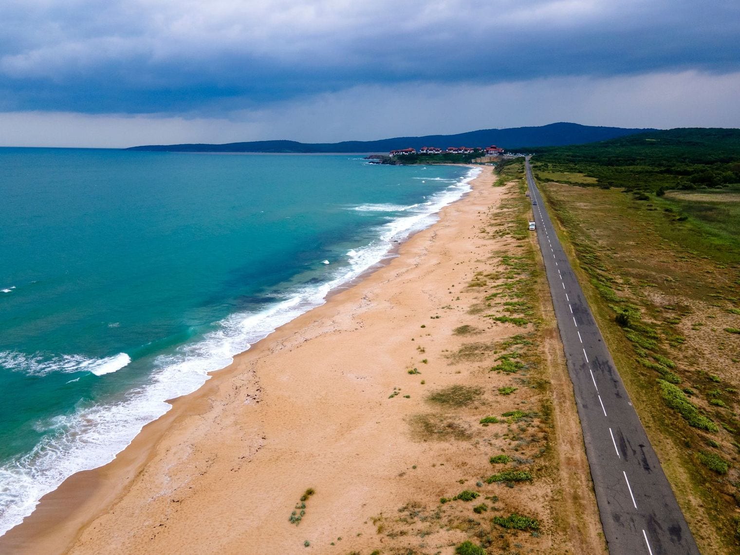 Aerial view of a long straight sandy beach running to the horizon beside a coastal road, with turquoise surf breaking along its full length