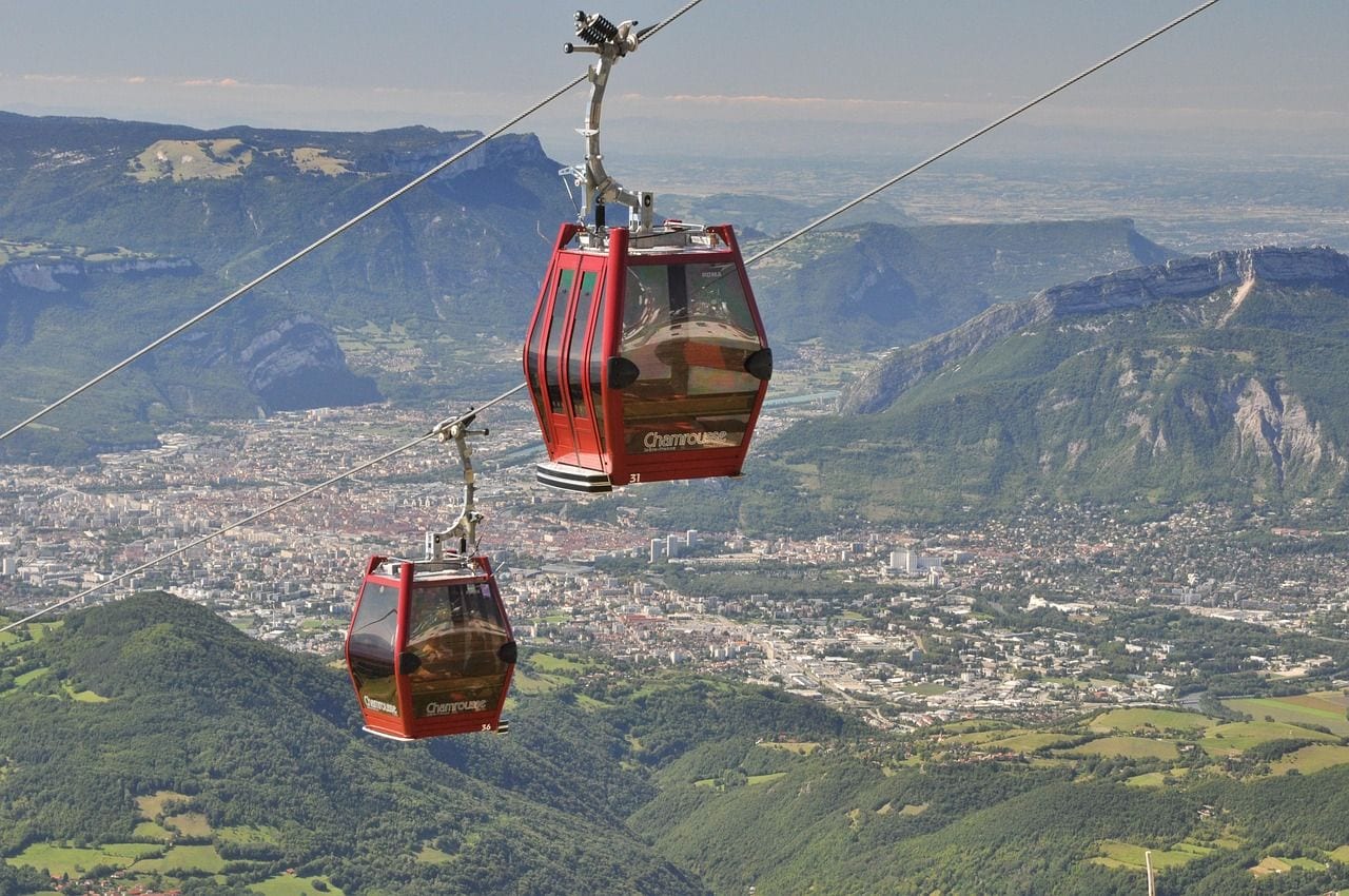 Two red gondola cabins riding a cable high above a green alpine valley, with a city spread across the plain far below