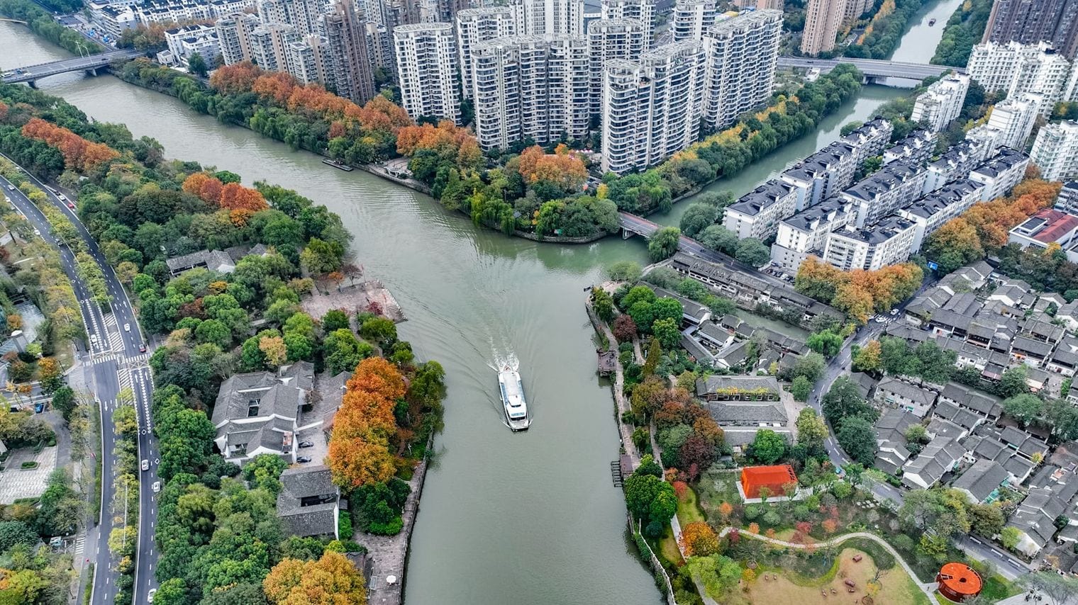 Aerial view of a passenger boat cruising a wide canal junction, with grey-tiled courtyard houses and autumn trees lining both banks below modern apartment towers.