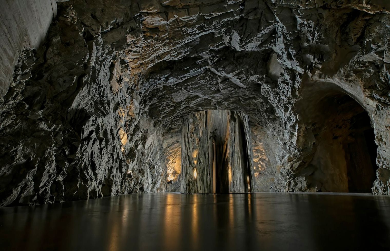 A lit passage deep in a cave, its rough rock walls and arched opening reflected in still water across the chamber floor