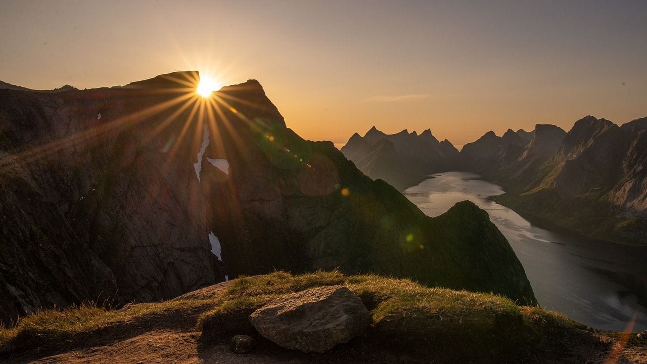 A low sun bursting over a dark mountain ridge above a narrow fjord, its water winding away between steep walls.