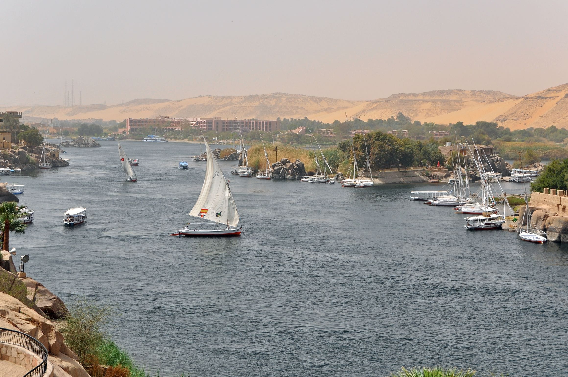 A white-sailed felucca crossing the Nile at Aswan, Egypt, with moored boats, granite rocks and golden desert dunes on the far bank