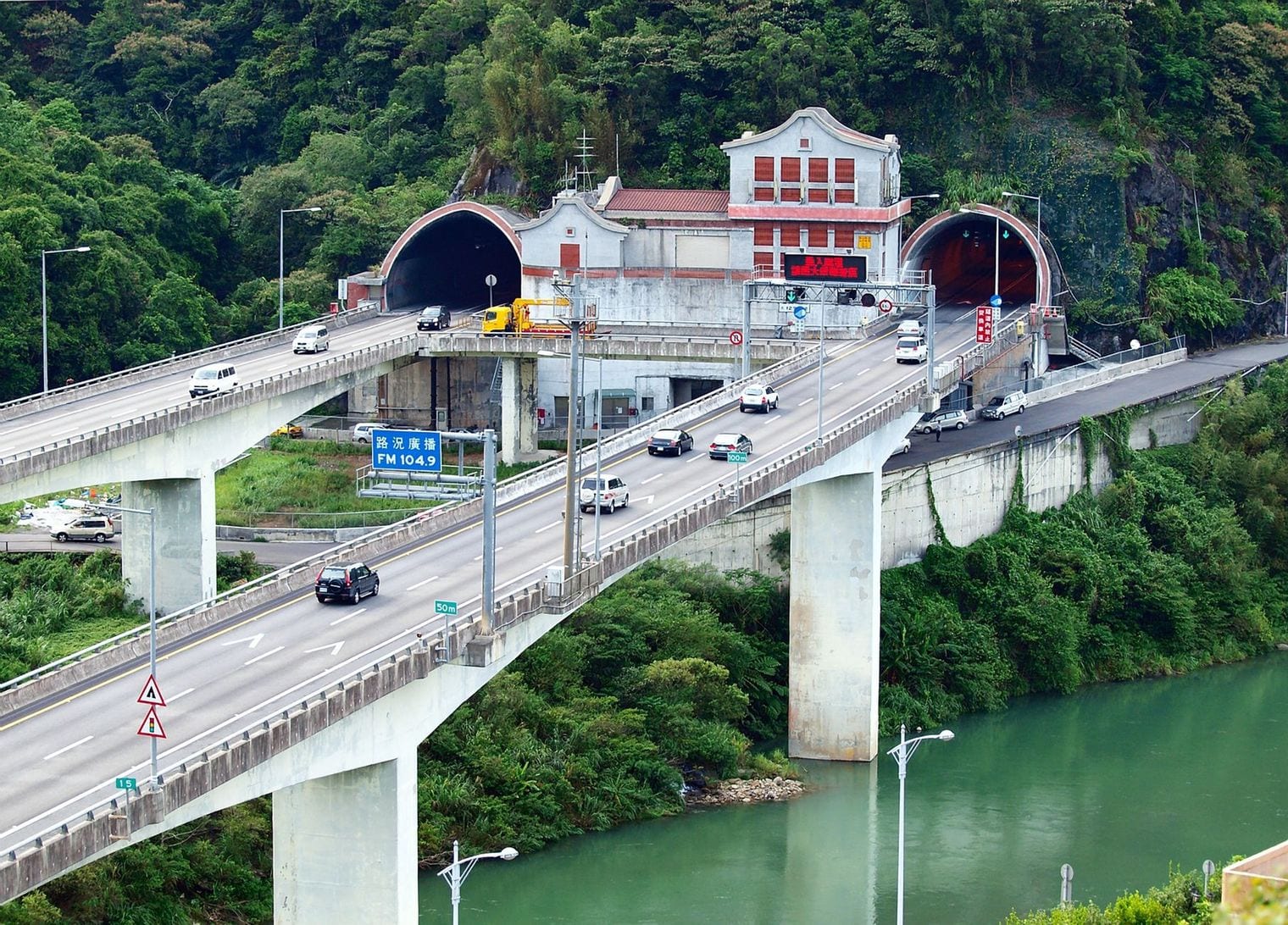 Twin arched portals of a mountain road tunnel set into a steep forested hillside, with an elevated motorway carried on concrete piers curving into them above a green river.