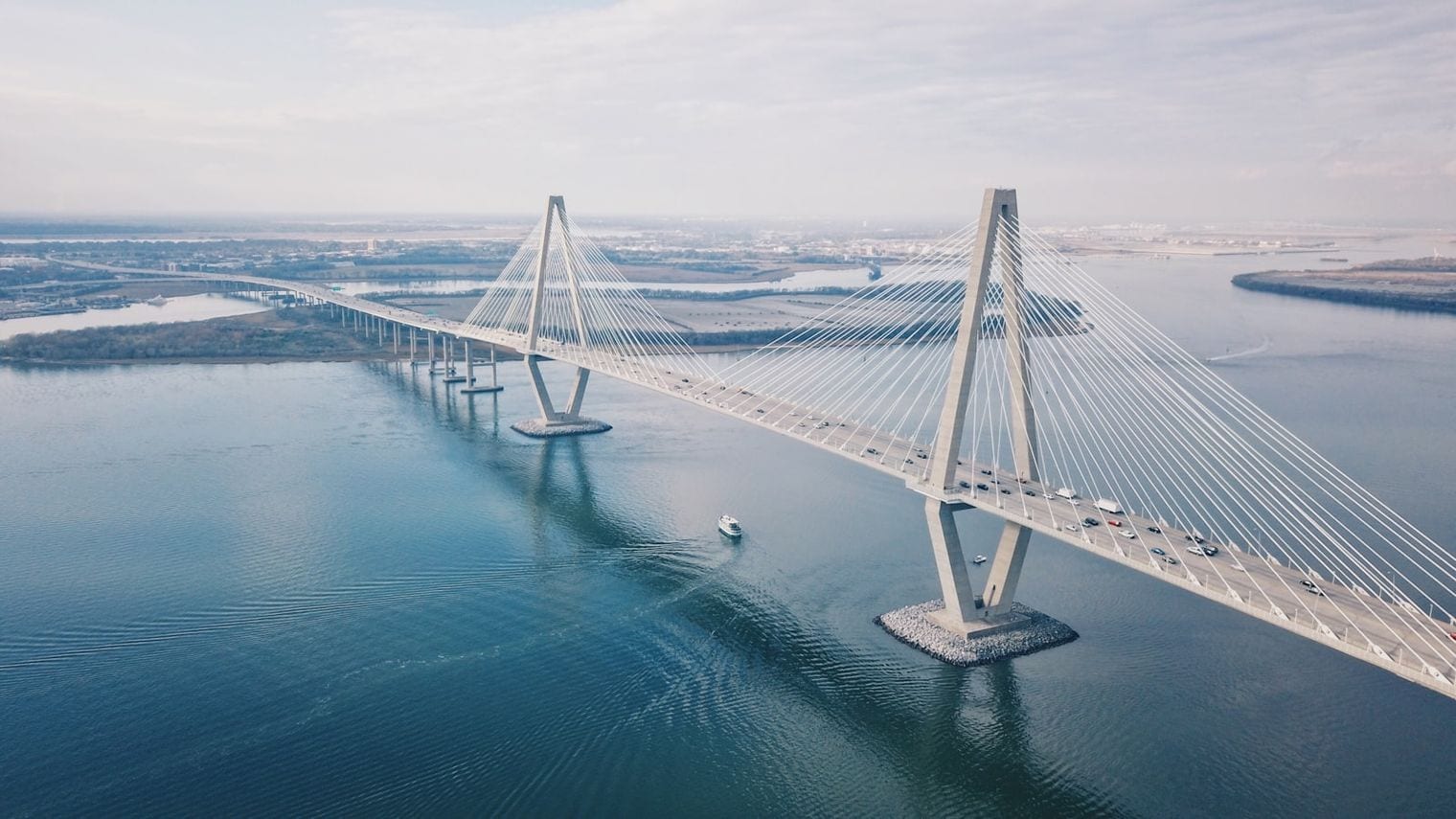 A cable-stayed bridge carrying a road low across open sea, its twin pylons silhouetted against a hazy sun.