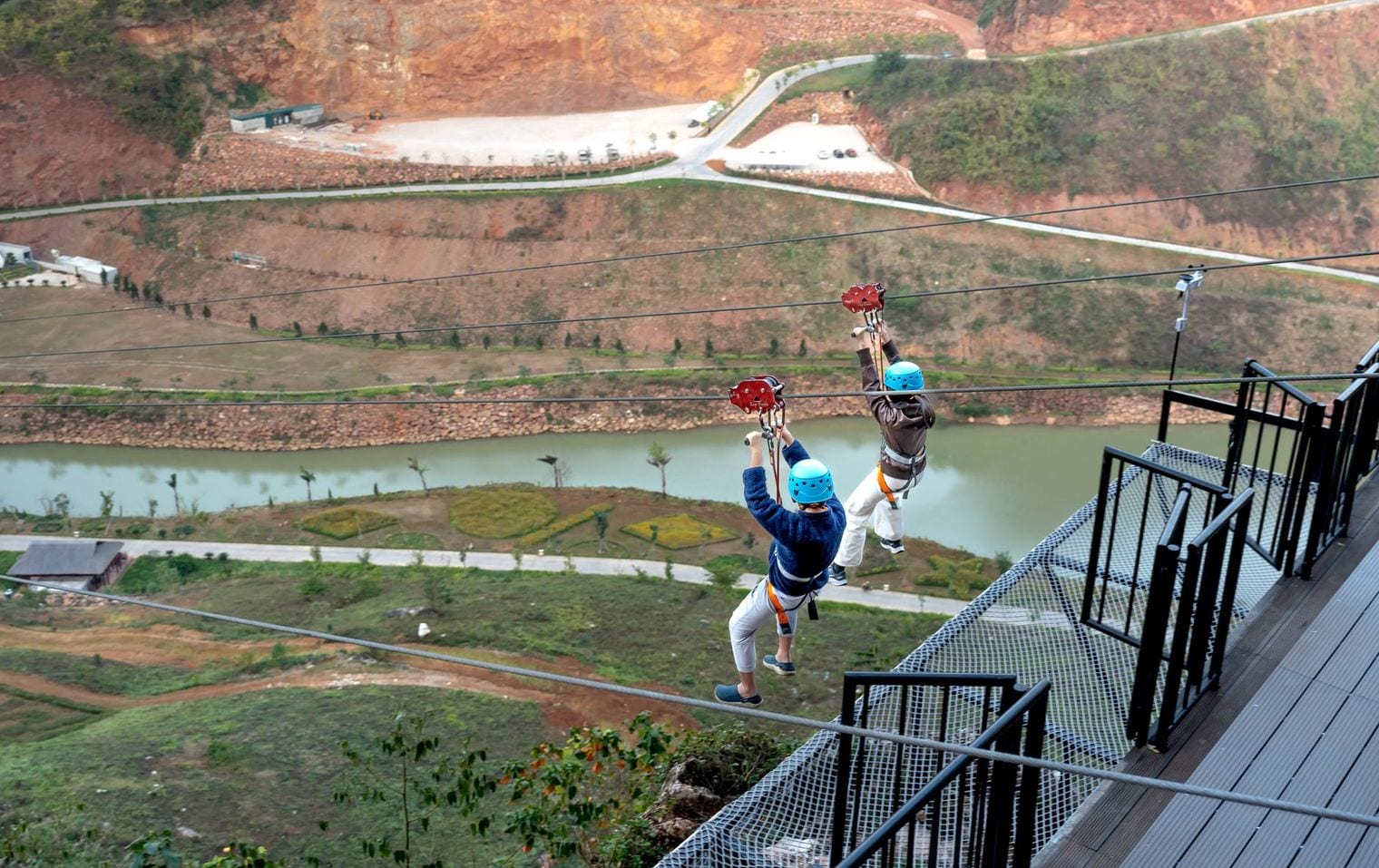 Two riders descending side by side on parallel zip-line cables above a river valley