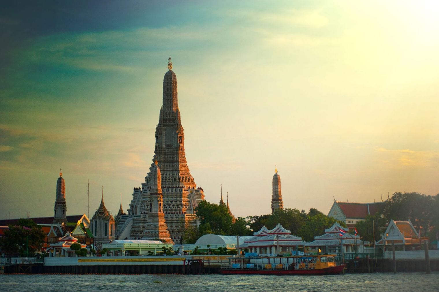 The riverside prang of Wat Arun catching low sunlight above the Chao Phraya in Bangkok, with passenger boats moored along the quay below
