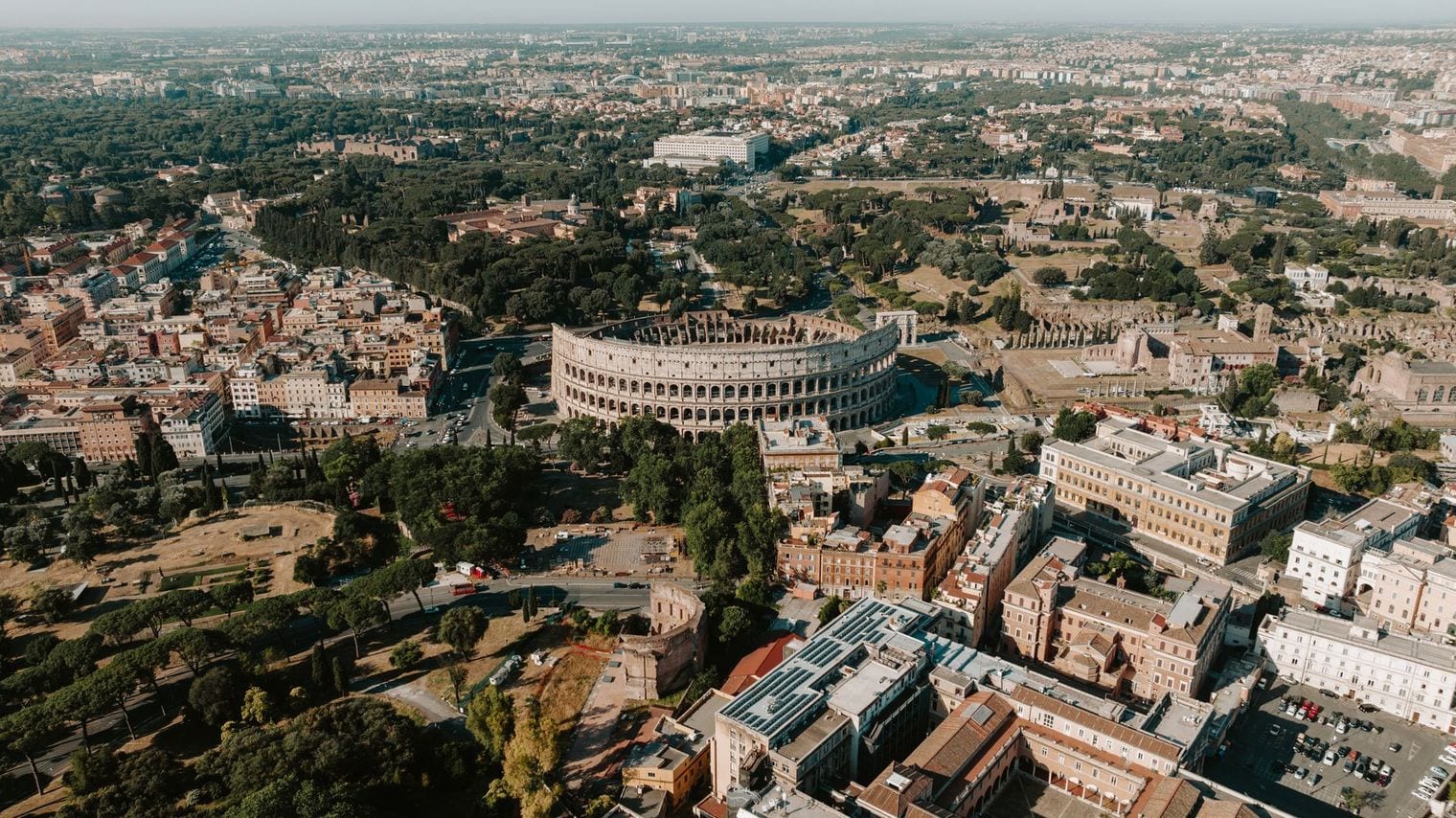 Aerial view across central Rome at golden hour, the Colosseum standing among streets and umbrella pines with the city stretching to the hills beyond