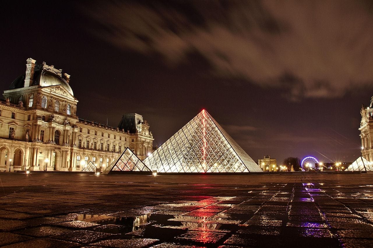 The Louvre's glass pyramid glowing at dusk in the Cour Napoleon, with the floodlit wings of the palace behind it and rain pooled across the courtyard.