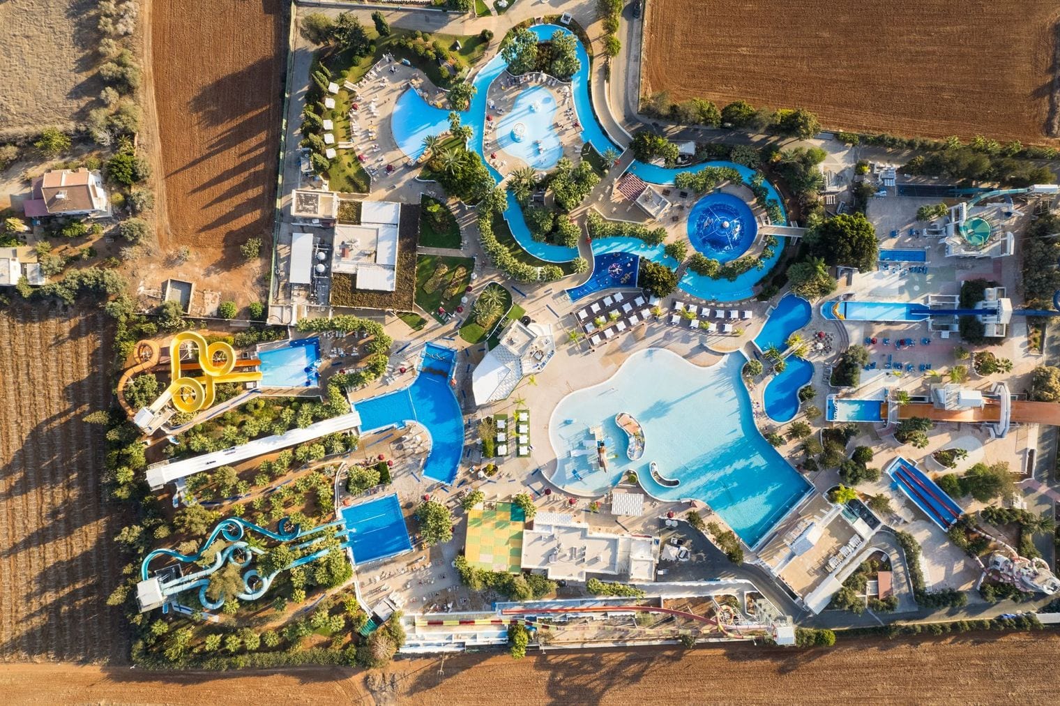 Aerial view of a water park, with looping slides, winding blue pools and sun terraces laid out among trees