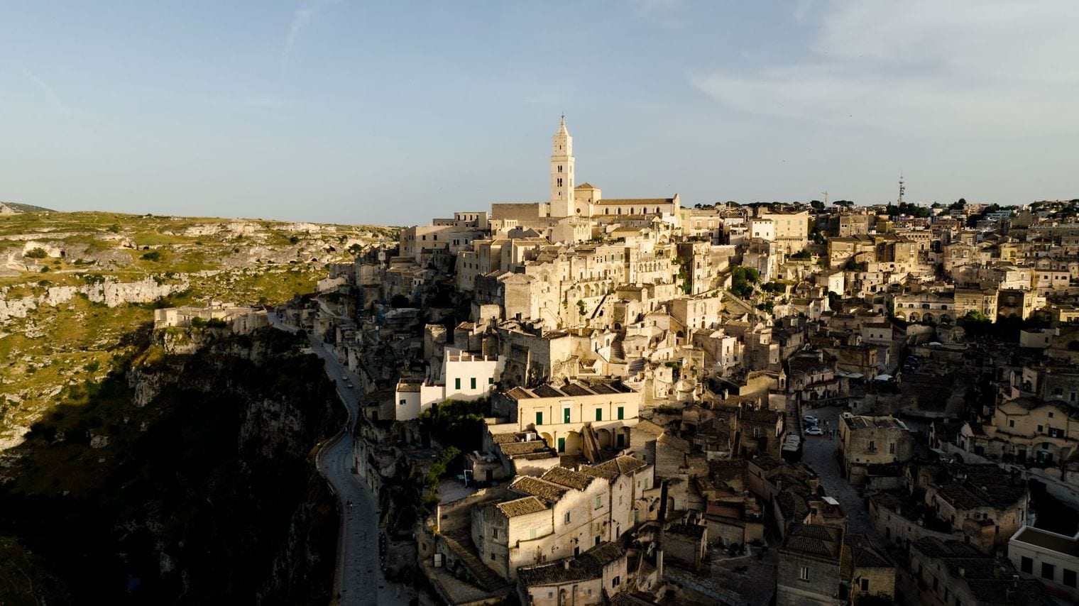 Aerial view of the Sassi of Matera at golden hour, stone houses and cave dwellings packed down the side of a limestone ravine with the cathedral bell tower rising above them