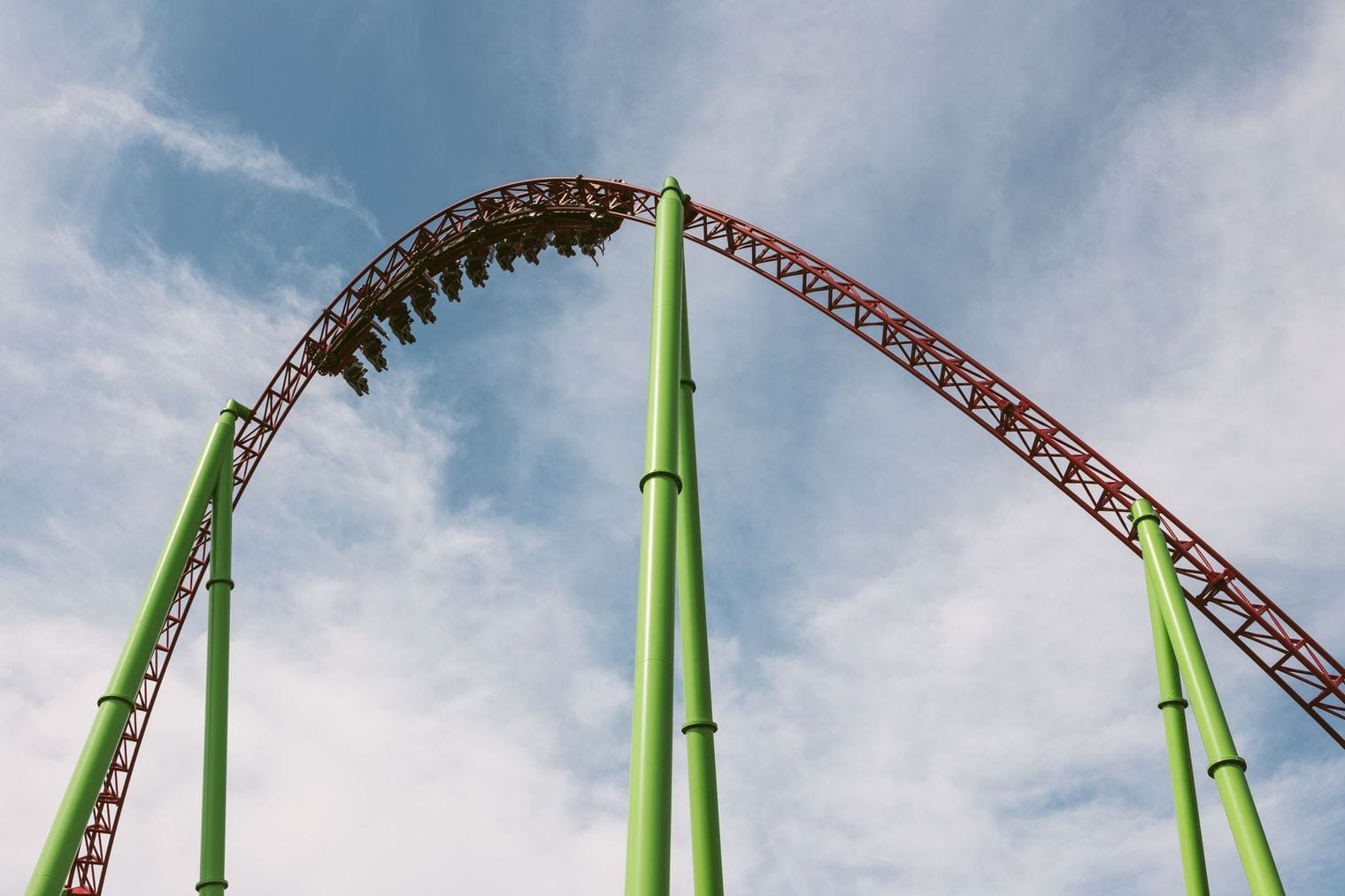 A red roller coaster track on bright green supports curving over the crest of an airtime hill against a blue sky, with a train of riders cresting the top