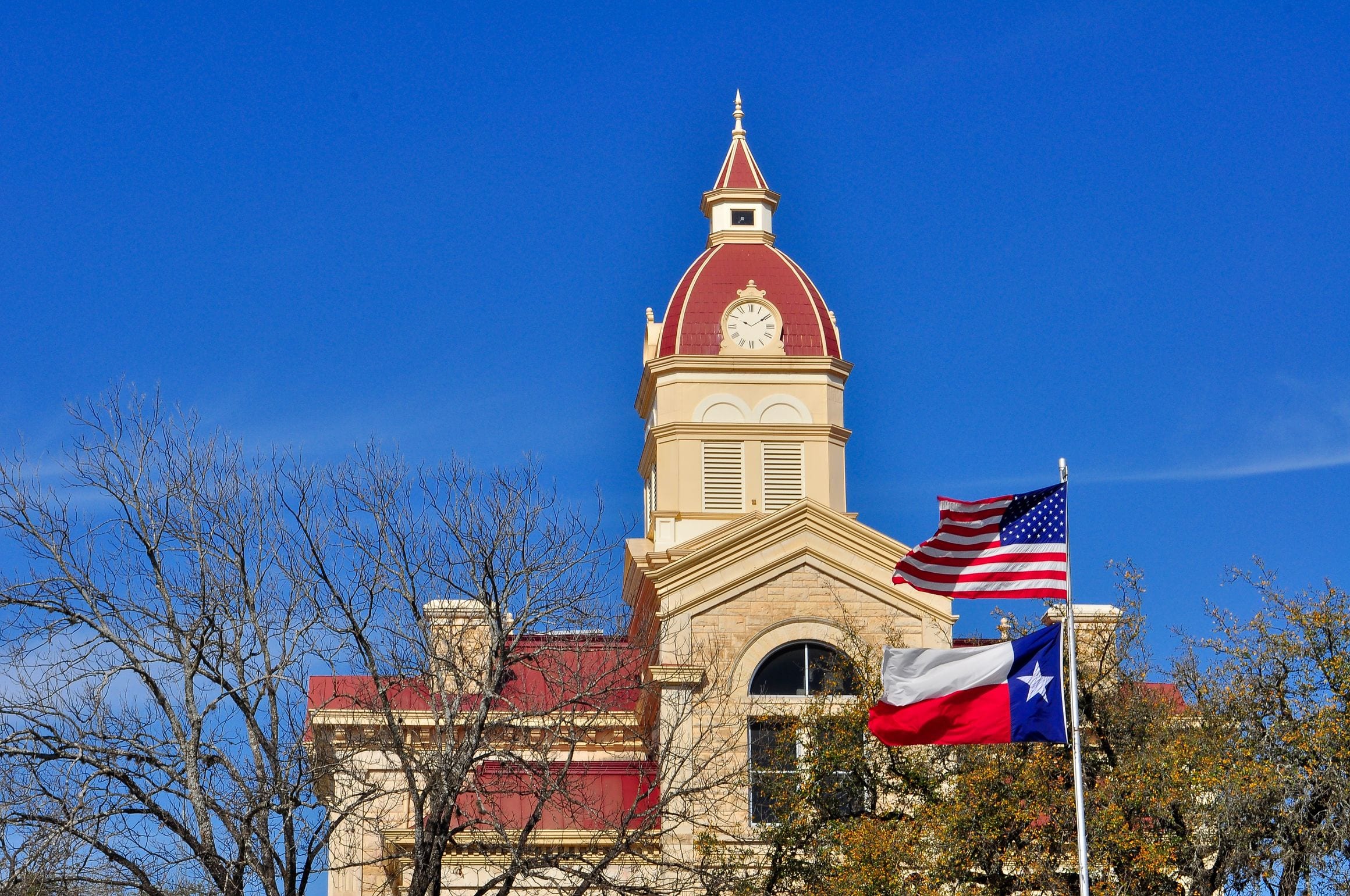 A small-town Texas courthouse in cream limestone with a red-roofed clock tower, the US and Texas flags flying beside it