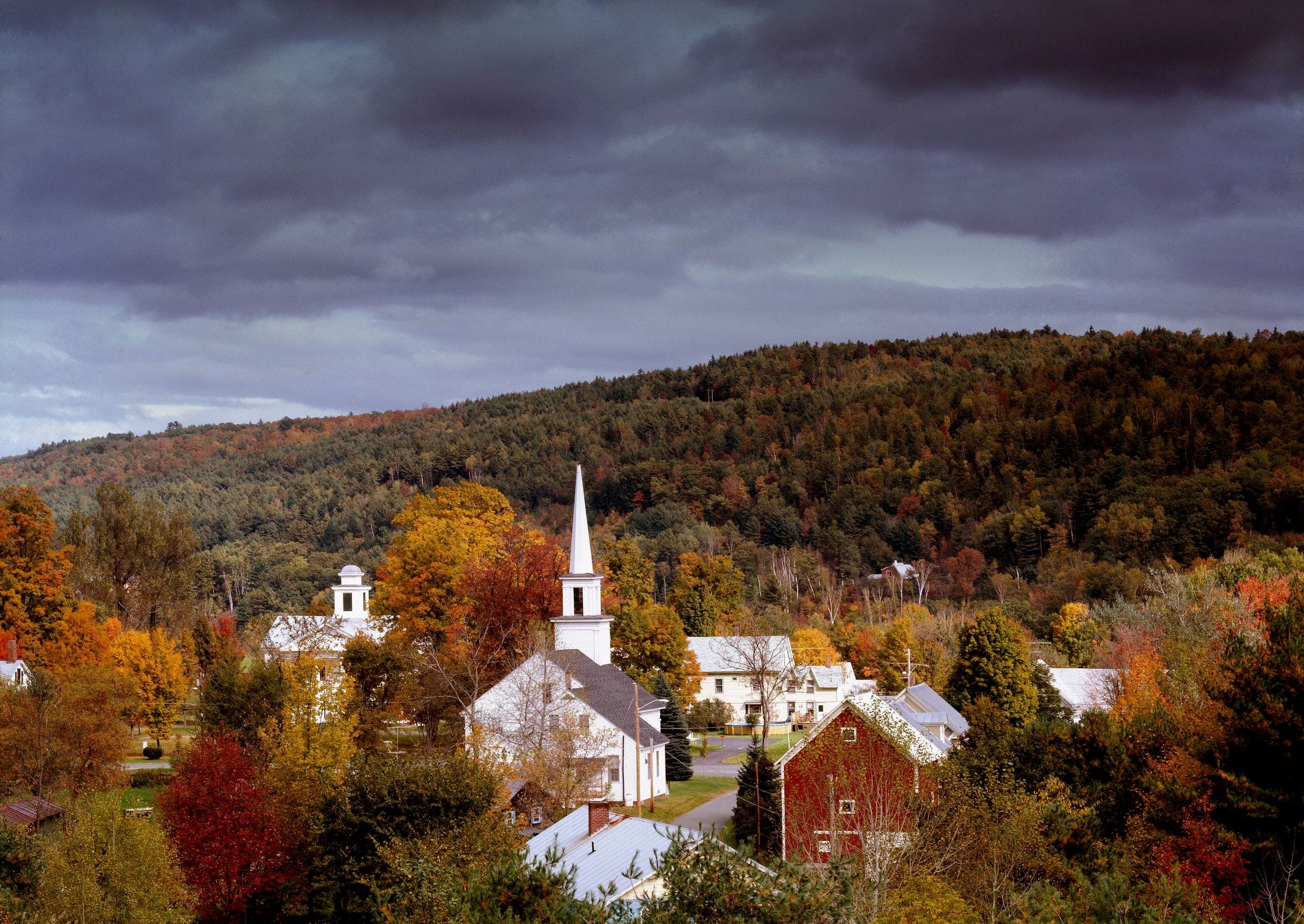 White church steeple rising over the village of Barnet, Vermont, among autumn foliage on a hillside beneath dark clouds