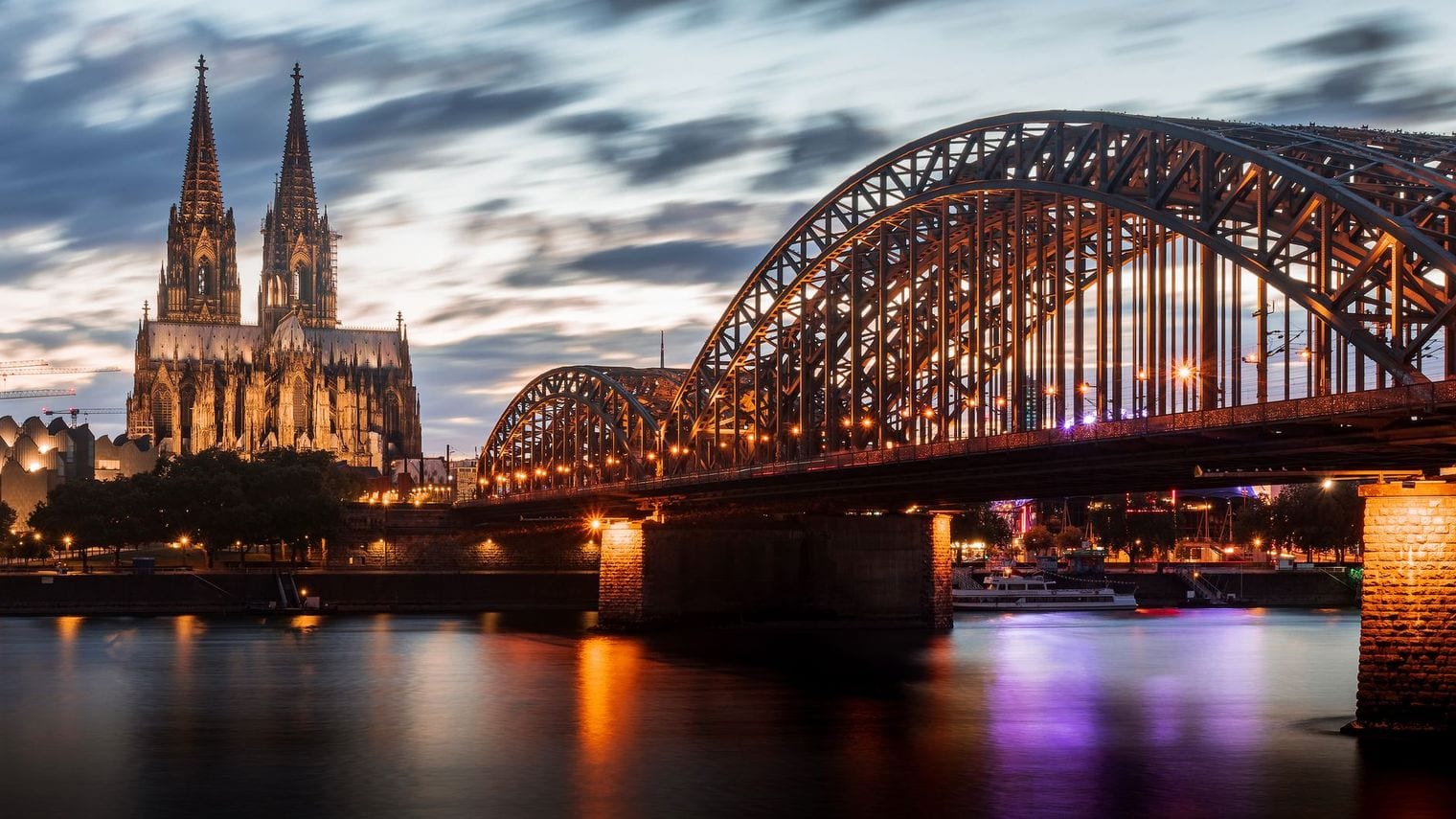 Cologne Cathedral floodlit at blue hour, its twin Gothic spires rising beside the steel arches of the Hohenzollern Bridge with the lights reflected in the Rhine