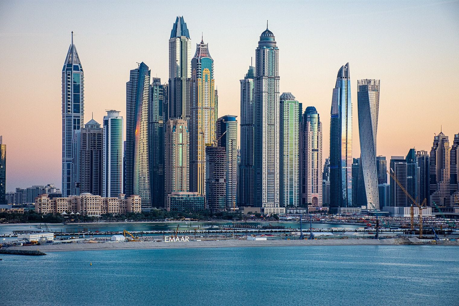 The Dubai Marina skyline at golden hour, a dense row of glass towers including the twisting Cayan Tower rising straight out of the water