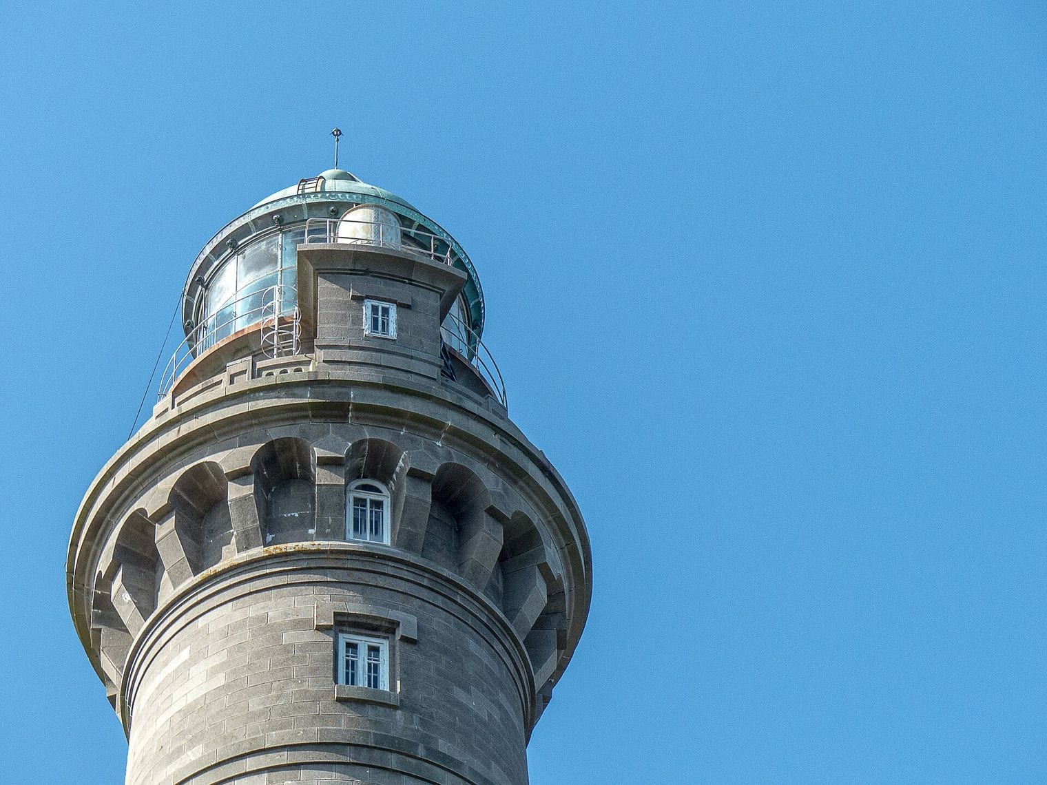 The corbelled granite gallery and copper-green lantern dome crowning a tall stone lighthouse, seen from below against a clear blue sky.