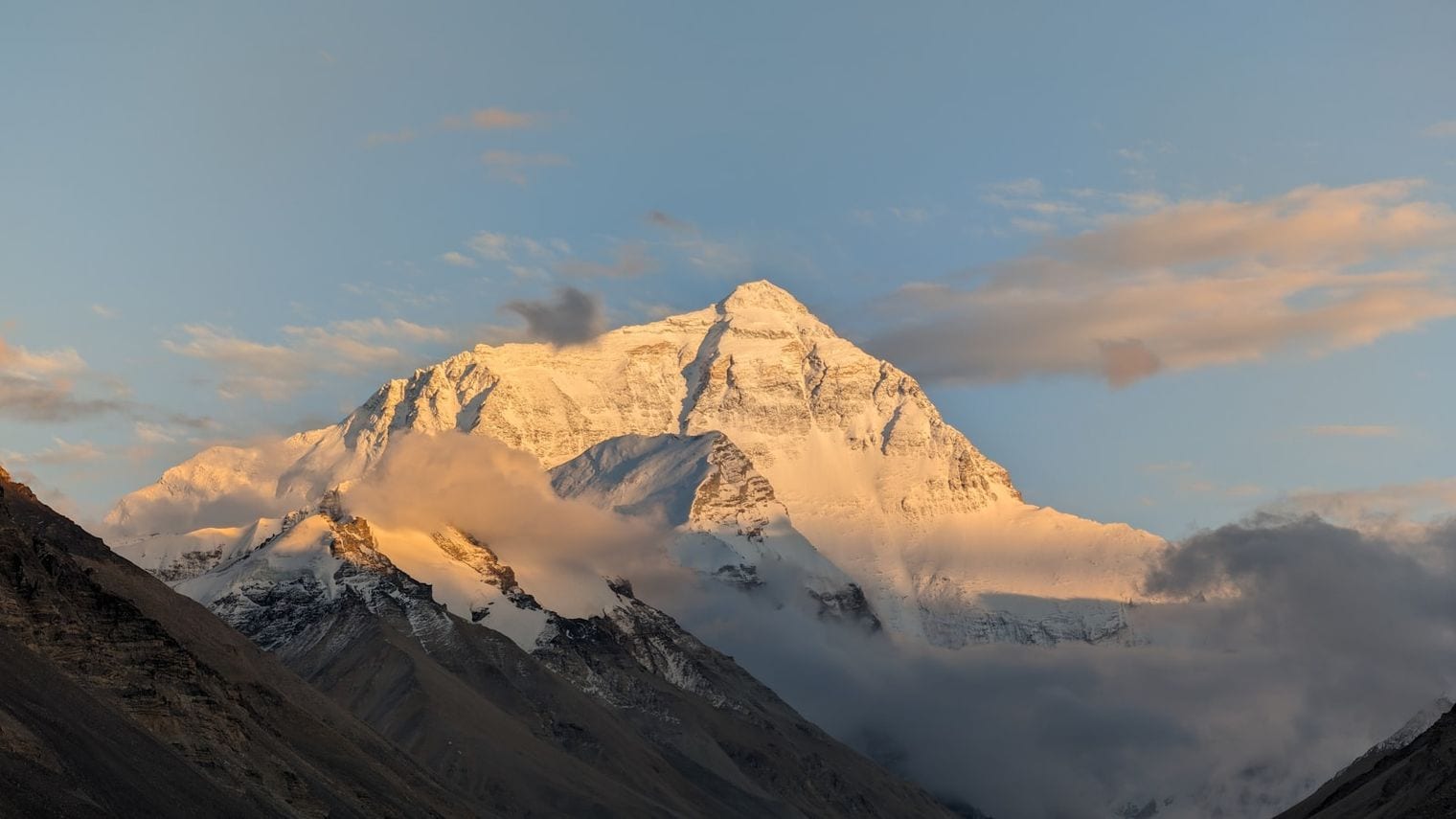 A snow-covered Himalayan summit lit by golden alpenglow, rising above a bank of cloud.