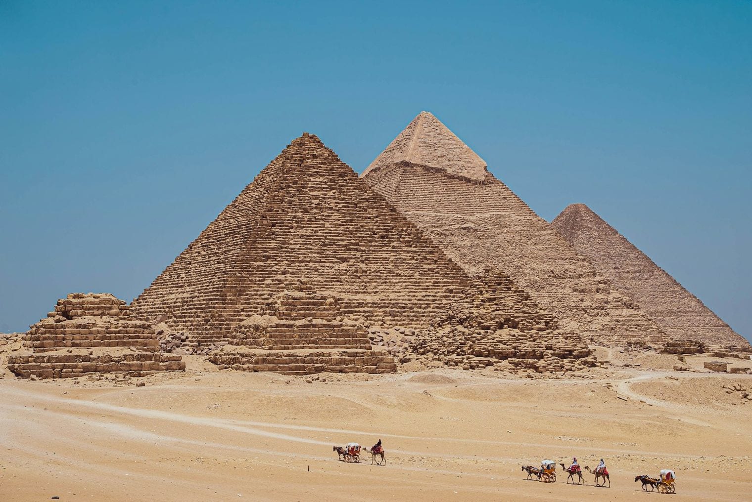The three main pyramids of Giza rising from open desert under a clear sky, with the smaller queens' pyramids and a group of camels in the foreground.