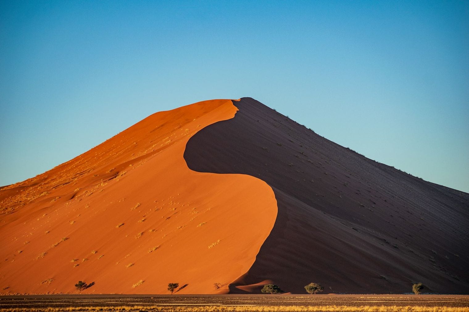 A lone figure walking the crest of a tall sand dune, footprints trailing along the ridge behind them, with rippled dunes rolling away to a hazy horizon.