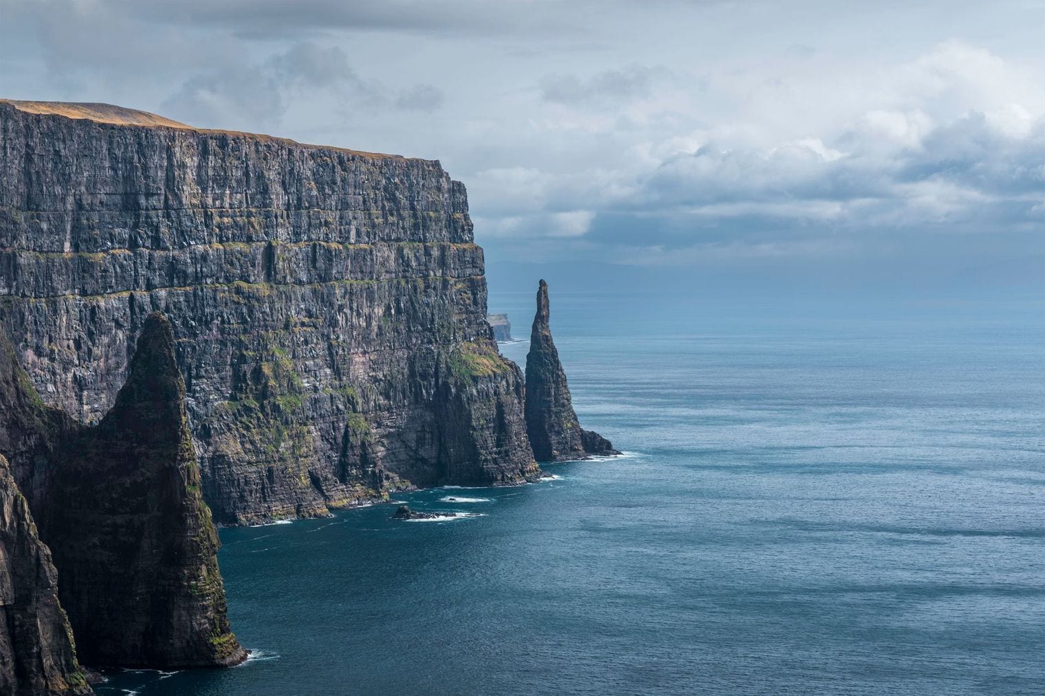 A towering wall of layered dark rock dropping sheer into deep blue ocean, with a slender sea stack standing just offshore beneath a bank of low cloud.
