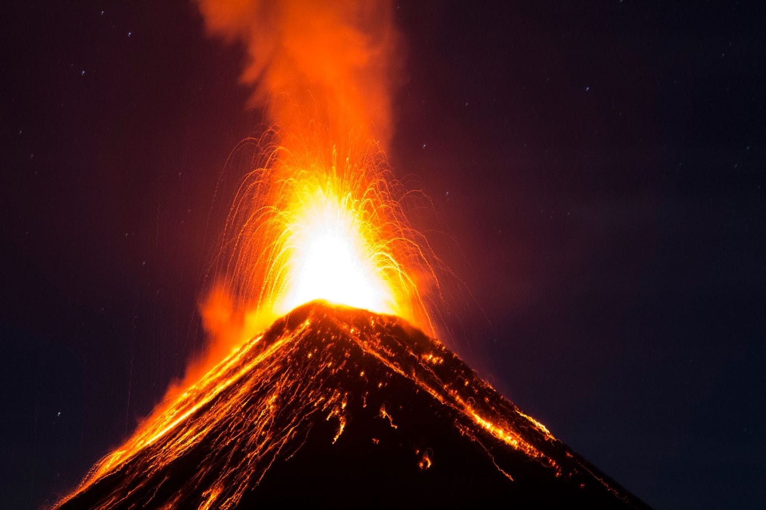 A volcano erupting at night, throwing a bright fountain of molten lava above its summit while glowing streams run down the dark flanks under a starry sky.