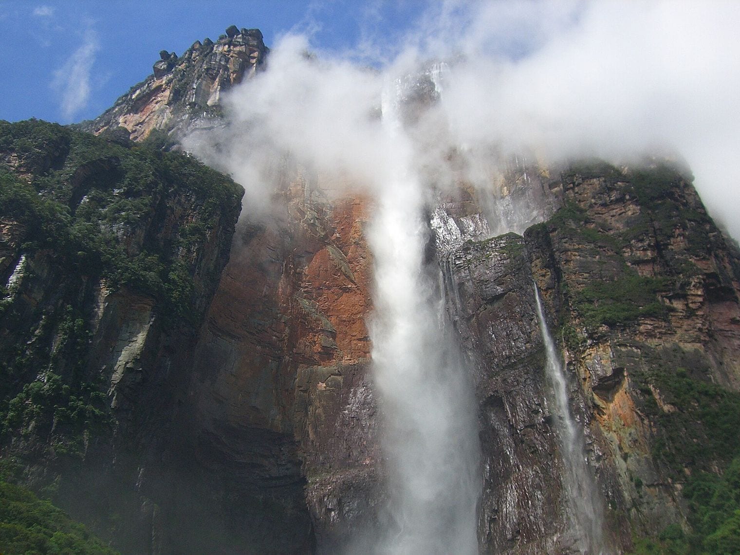 A single plume of water falling from the rim of a sheer ochre and grey tepui cliff, breaking into drifting mist against the rock face, with cloud across the summit and dark jungle crowding the foot of the wall.