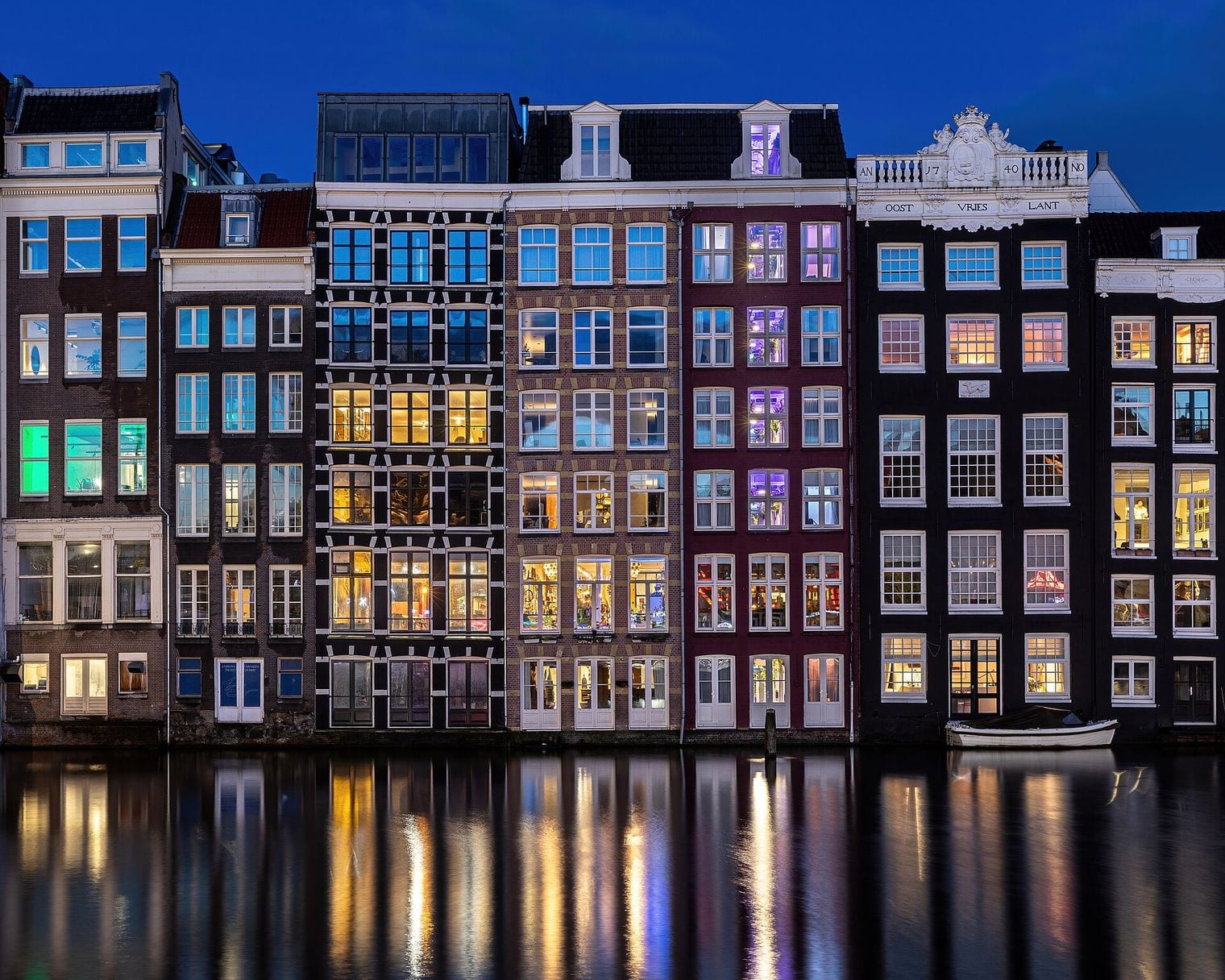Tall gabled canal houses lit from within at blue hour, reflected in the still water of the Damrak in Amsterdam.