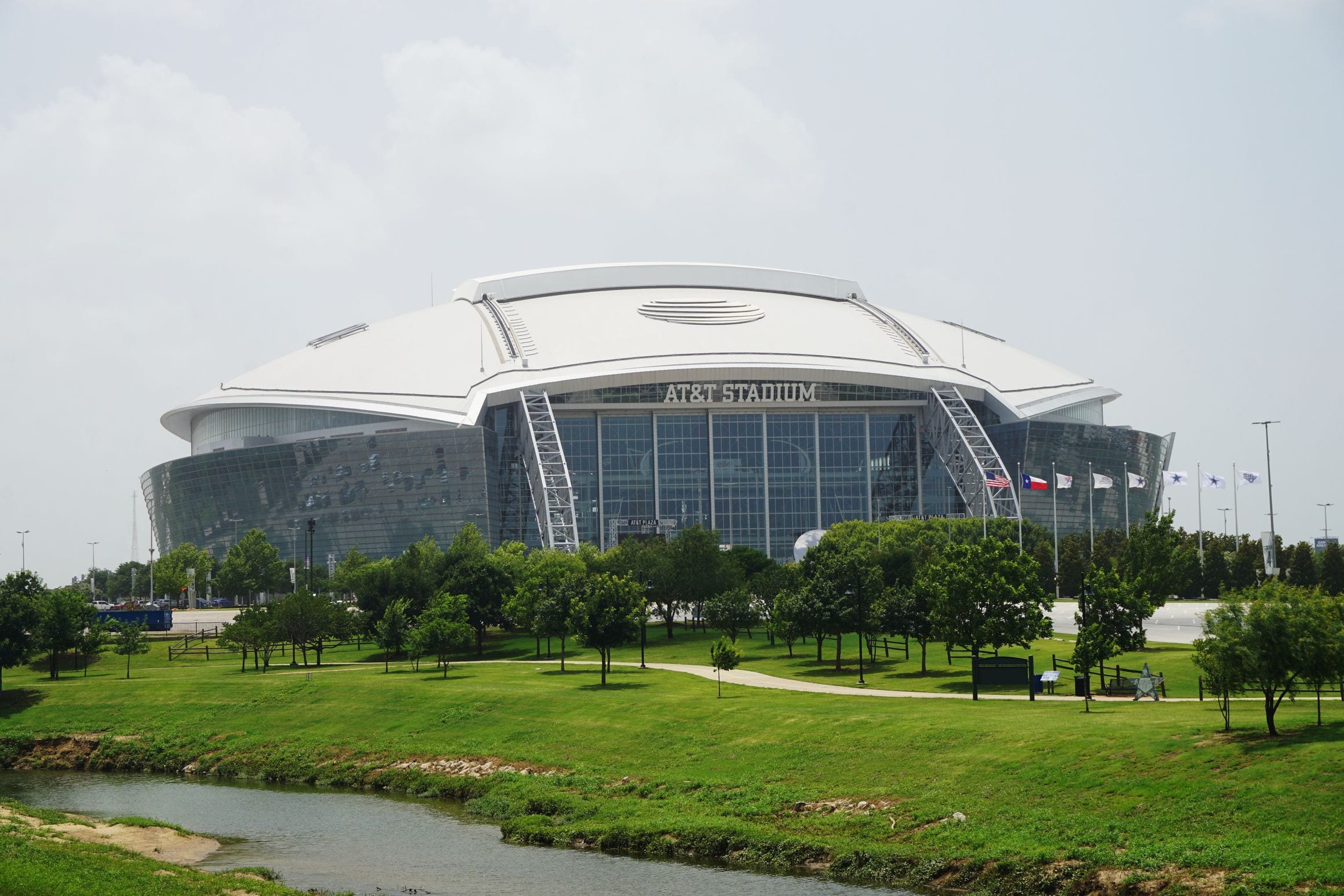 AT&T Stadium in Arlington, Texas, its domed roof and glass end wall rising behind a tree-lined park