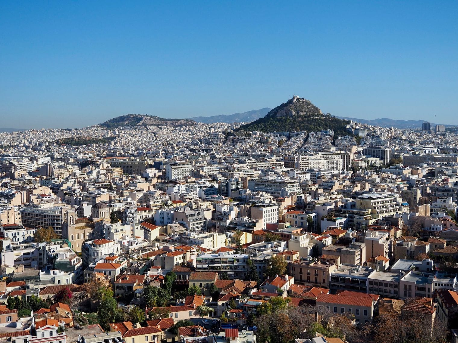 A daytime panorama across the white apartment blocks and red-tiled rooftops of central Athens, with the pine-covered cone of Mount Lycabettus rising behind the city