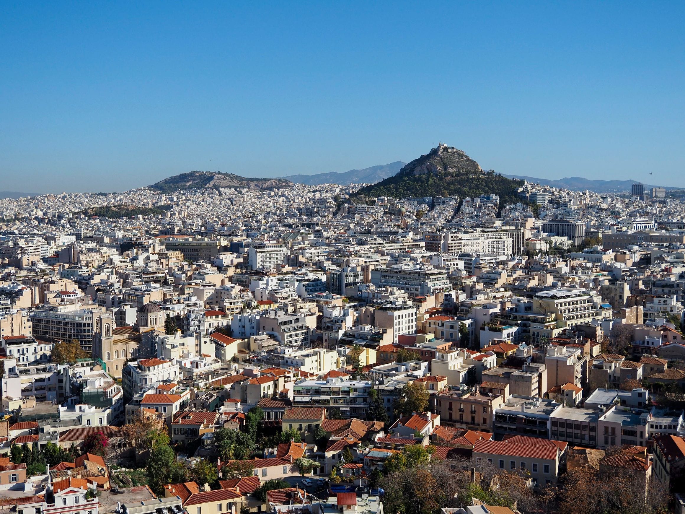 A daytime panorama across the white apartment blocks and red-tiled rooftops of central Athens, with the pine-covered cone of Mount Lycabettus rising behind the city