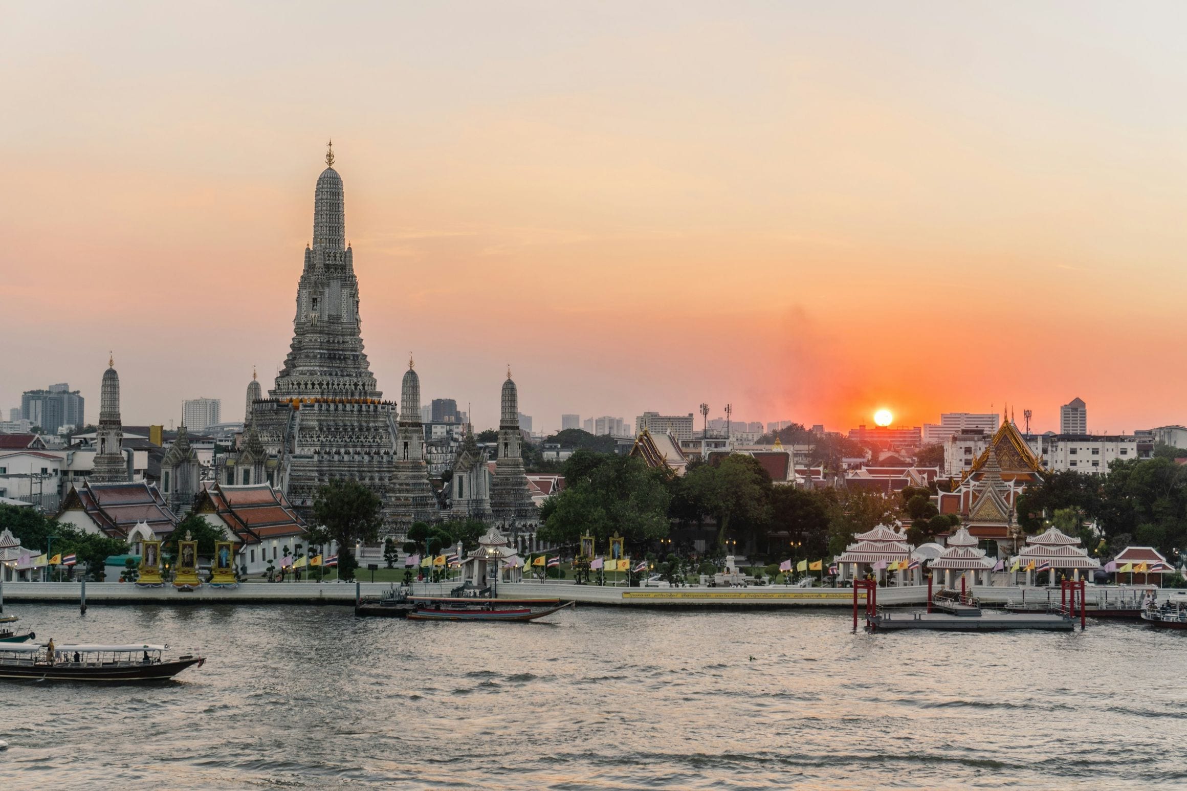 The porcelain-covered prang of Wat Arun silhouetted against an orange sunset across the Chao Phraya River, with longtail boats moored along the far bank