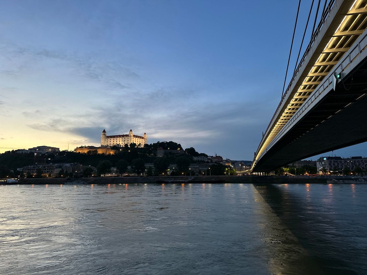 Bratislava Castle floodlit on its bluff above the Danube at dusk, with the cable-stayed SNP Bridge crossing the river in the foreground.