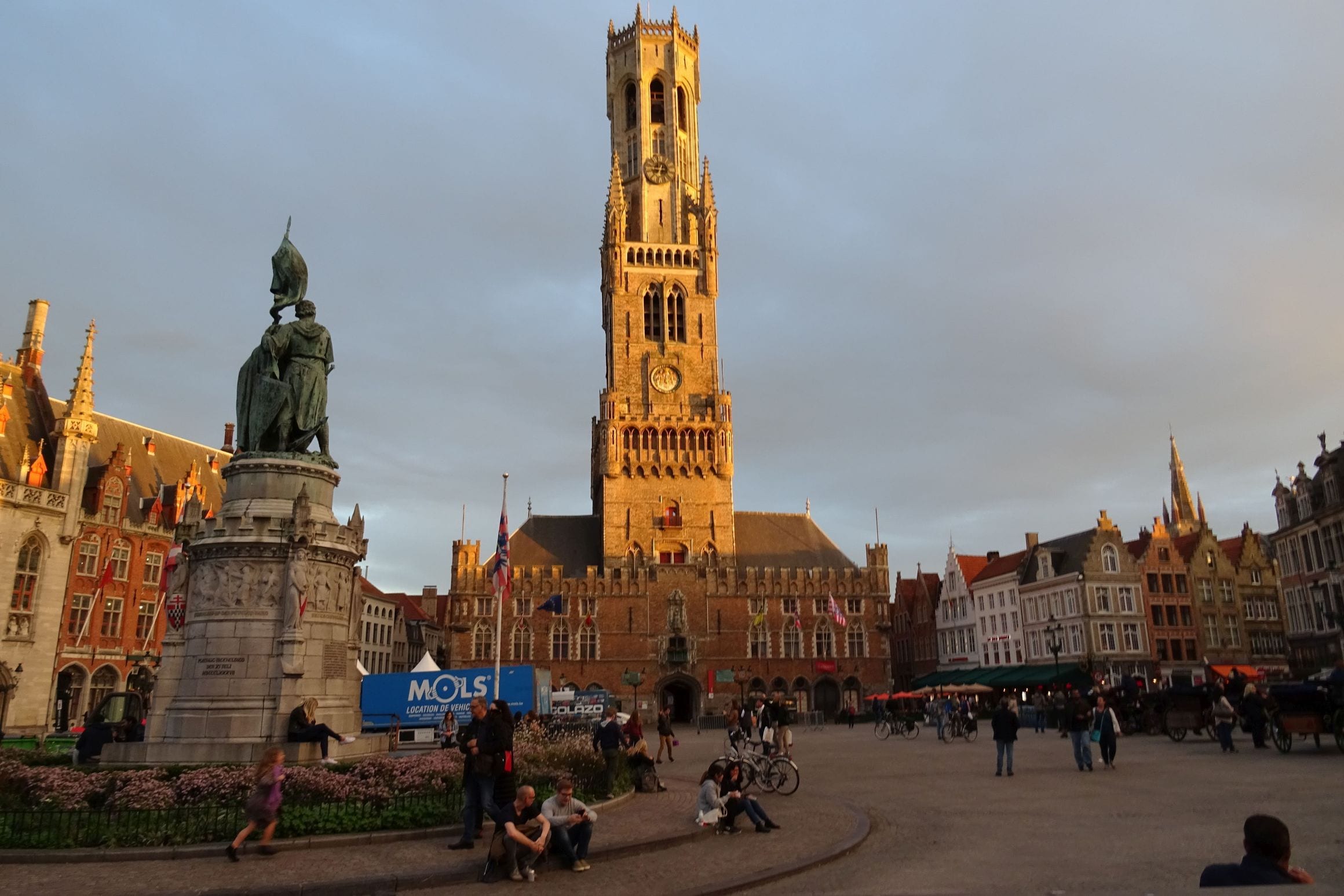 Golden-hour view across the Markt in Bruges, with the Belfry tower rising above the guild houses and the Jan Breydel and Pieter de Coninck statue in the square